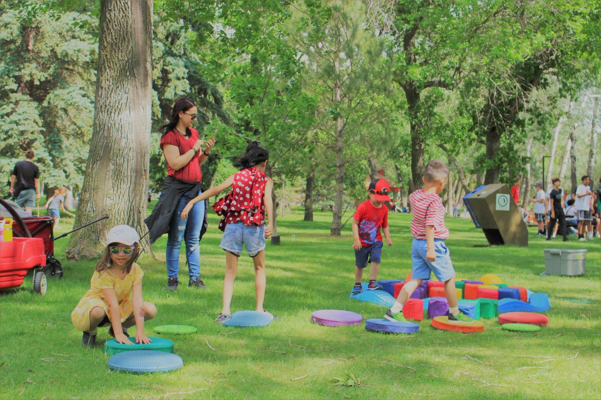 Children play on colourful foam pads in a grassy park while adults stand nearby.