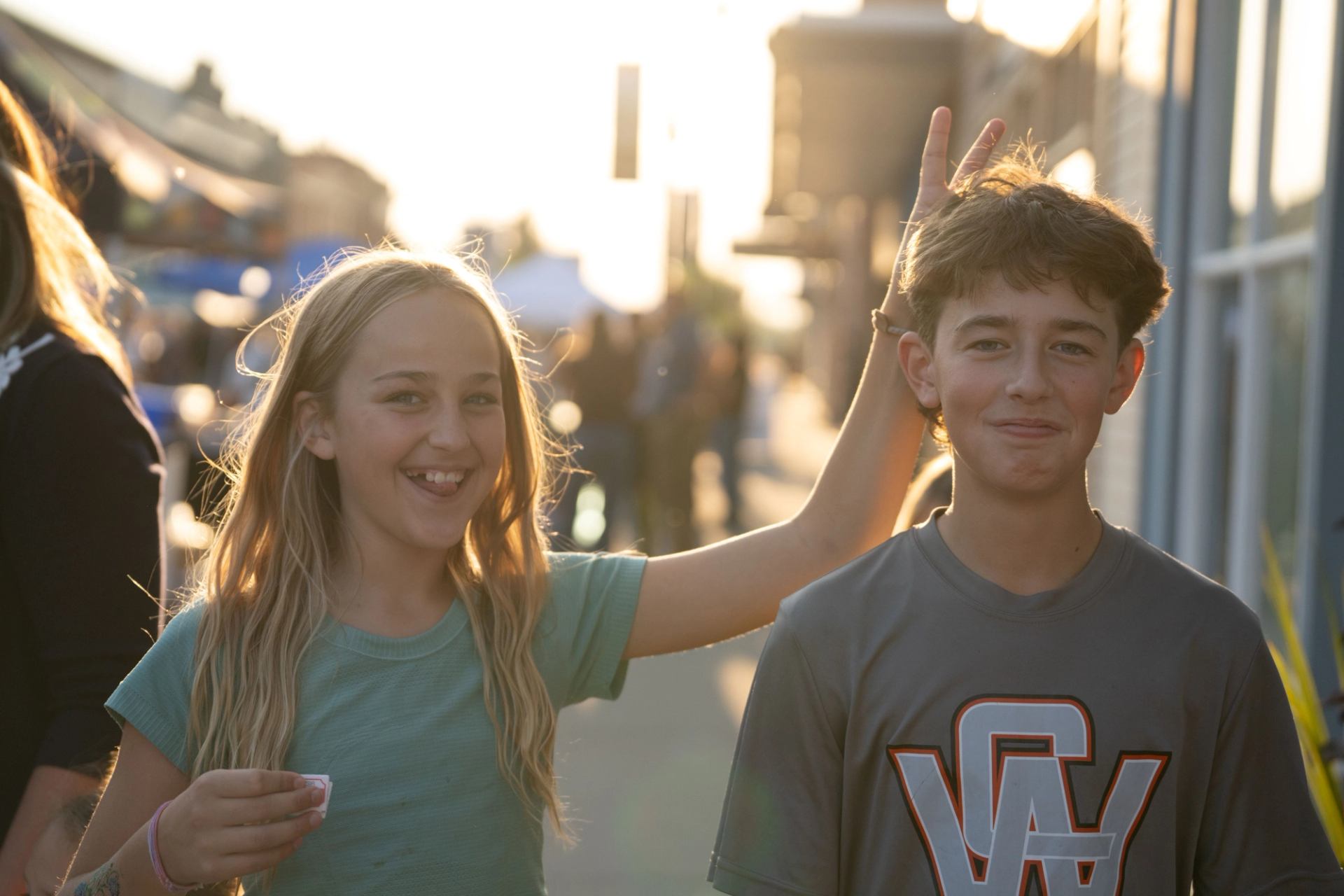 Two kids stand on a sunlit street, one holding ice cream and making bunny ears.