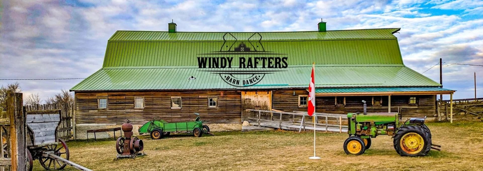 A large rustic wooden barn with a green roof displays a sign reading Windy Rafters Barn Dance. A vin