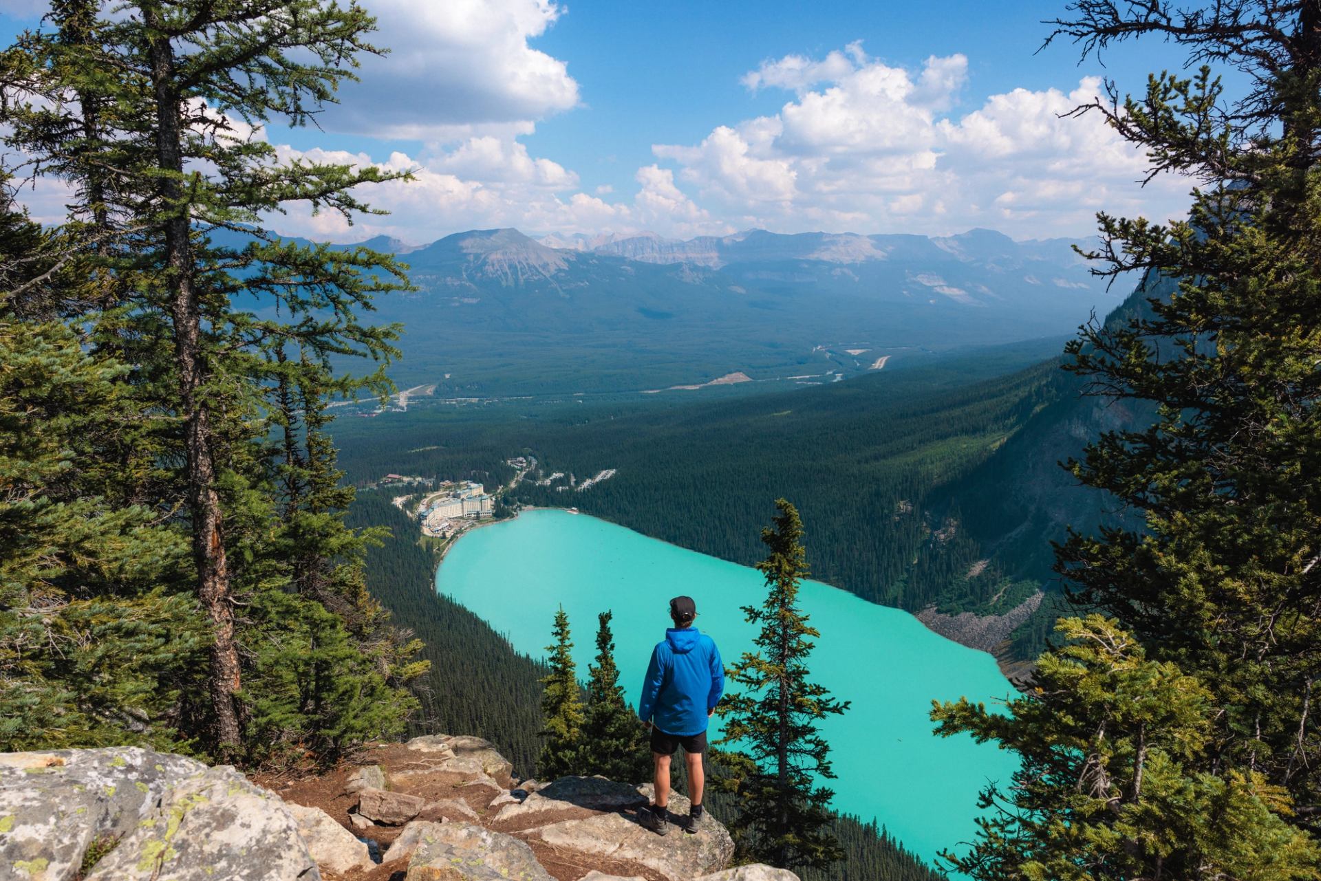 Hiker in blue jacket overlooks turquoise lake, forested valley and distant mountain range.