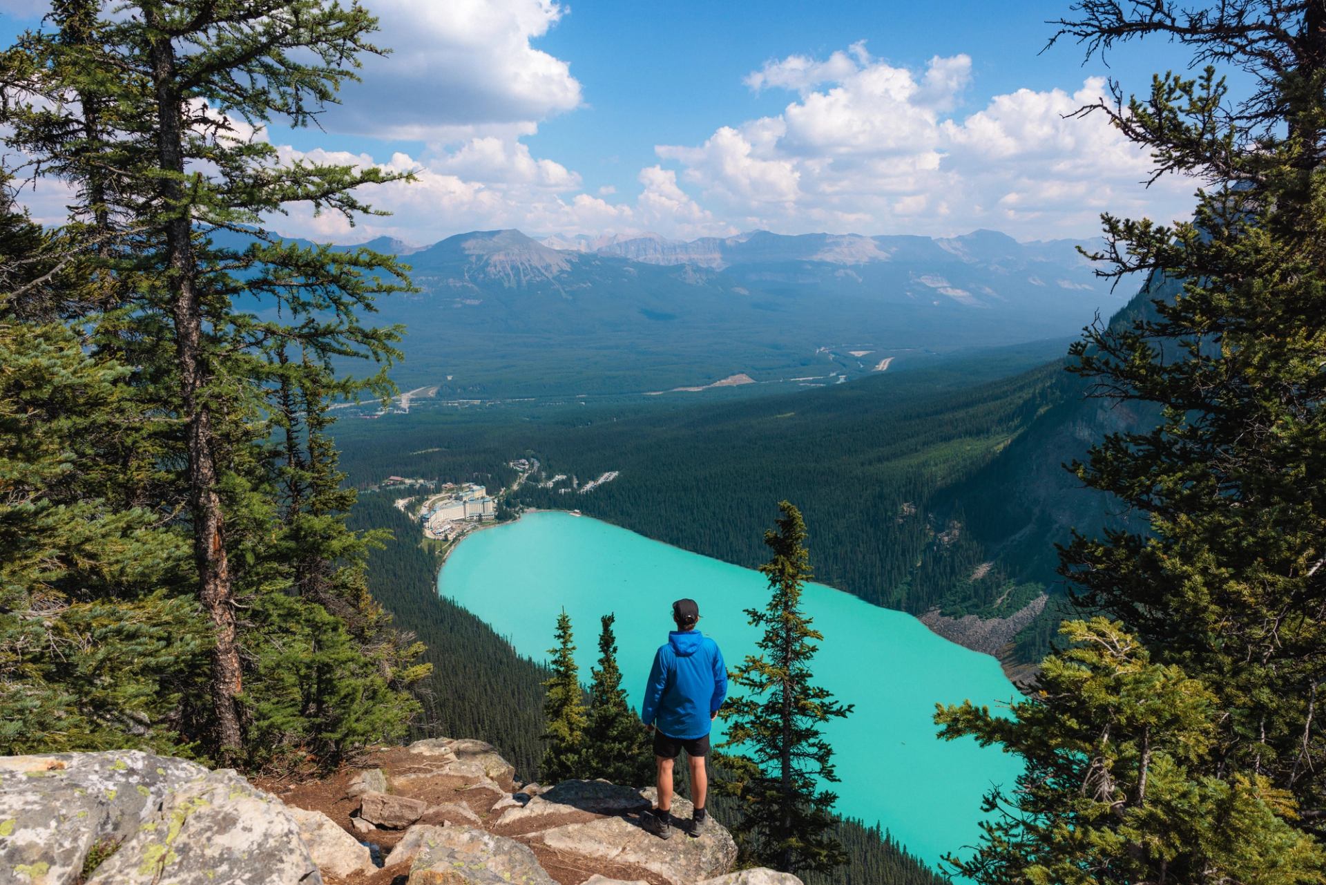 Hiker in blue jacket overlooks turquoise lake, forested valley and distant mountain range.