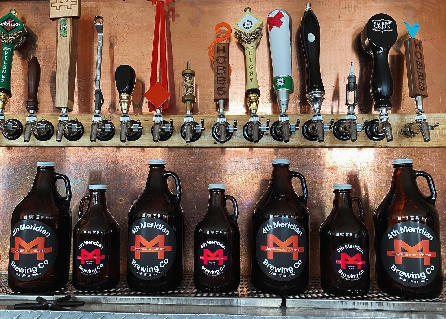 Beer taps with assorted handles above 4th Meridian Brewing Co growlers on metal shelf.
