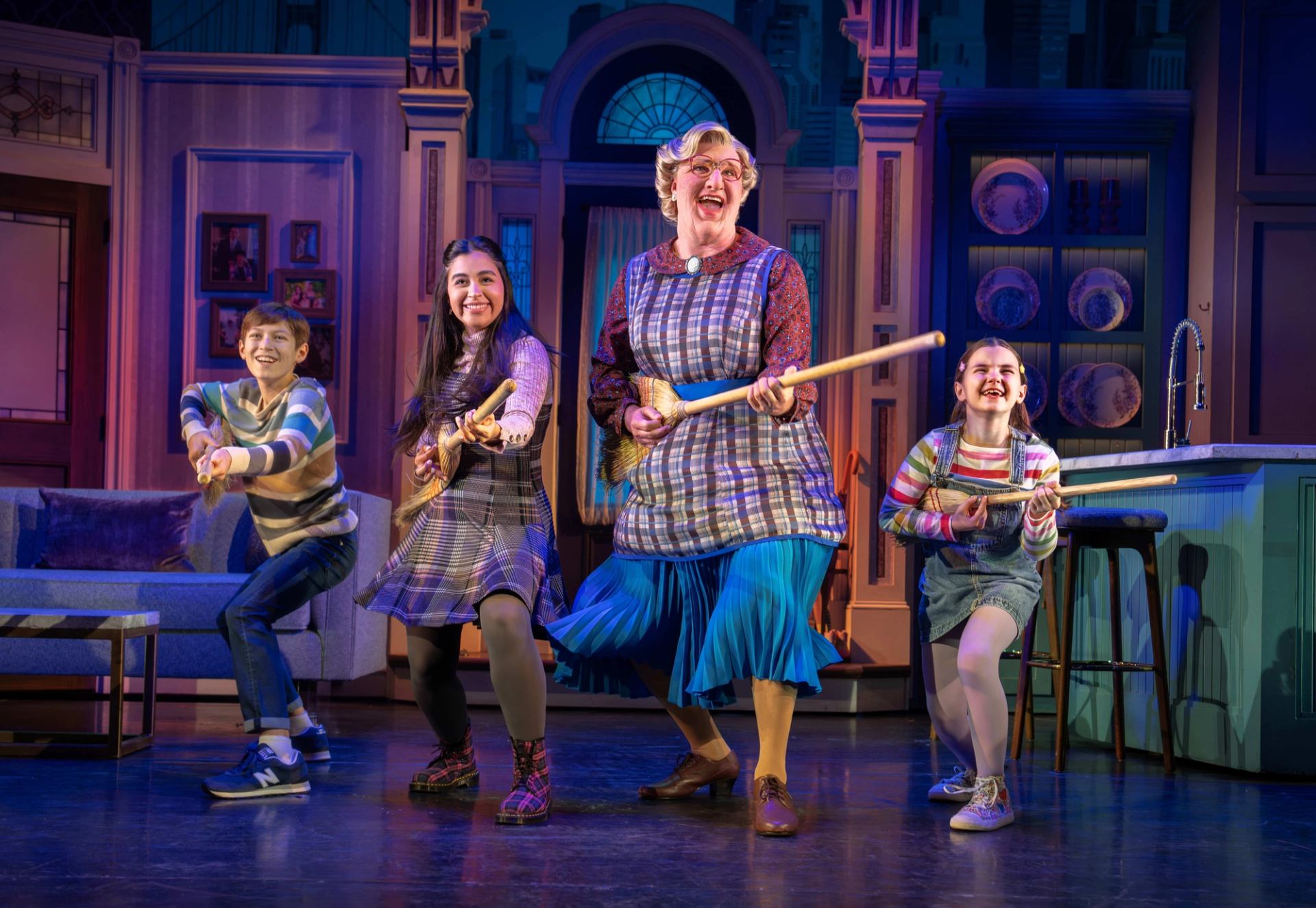 Stage performers in colourful costumes pretend to play guitars with brooms in a kitchen set.