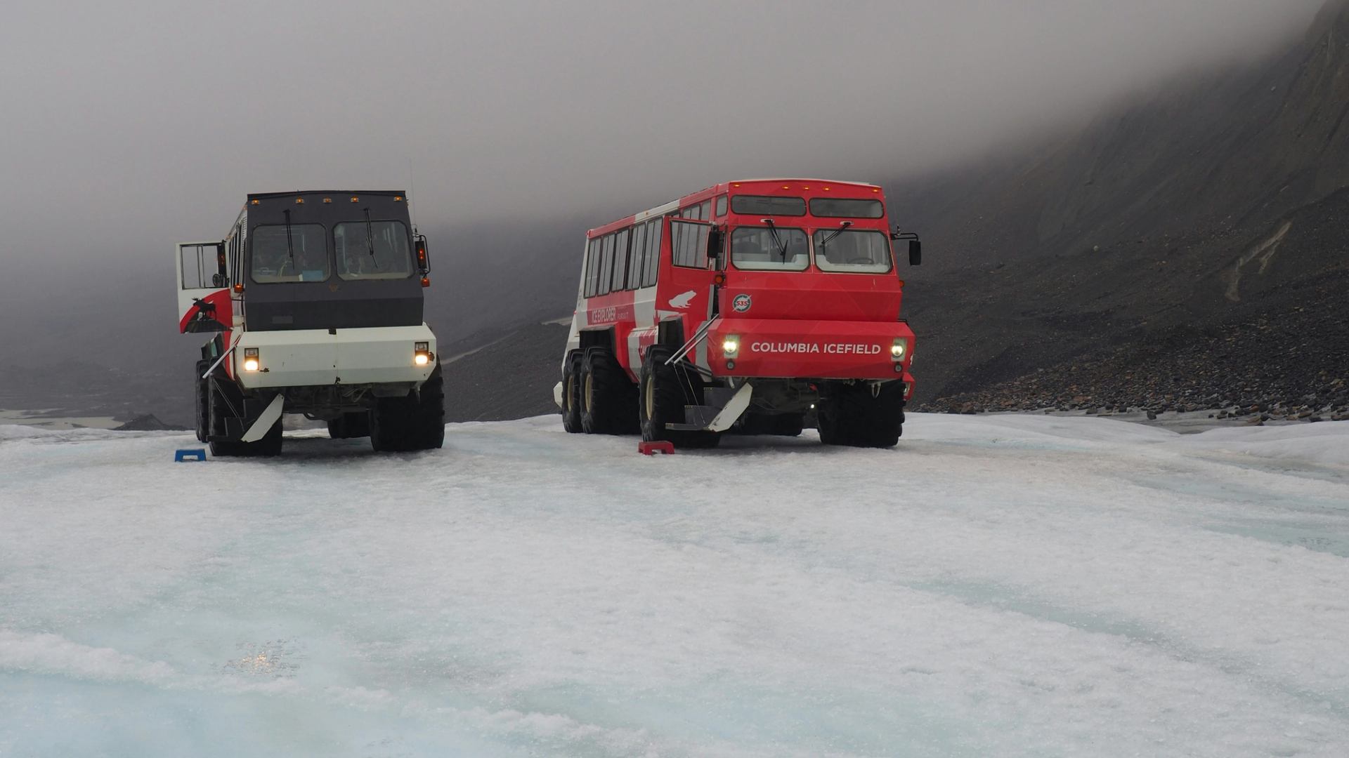 Two Columbia Icefield tour buses parked on a glacier beneath low clouds and rocky slopes.