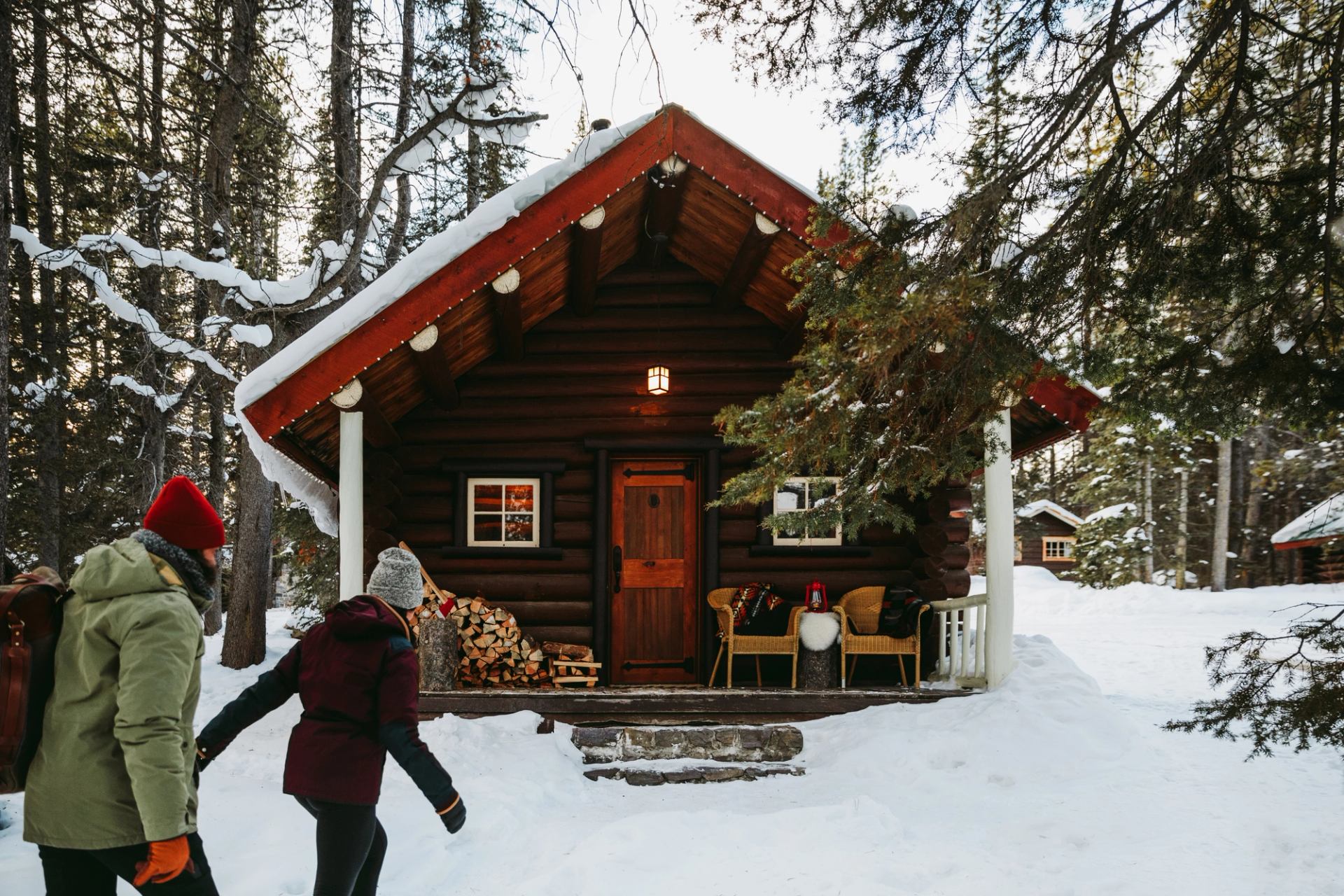 two people approach cabin through snow, surrounded by trees and warm porch lights.