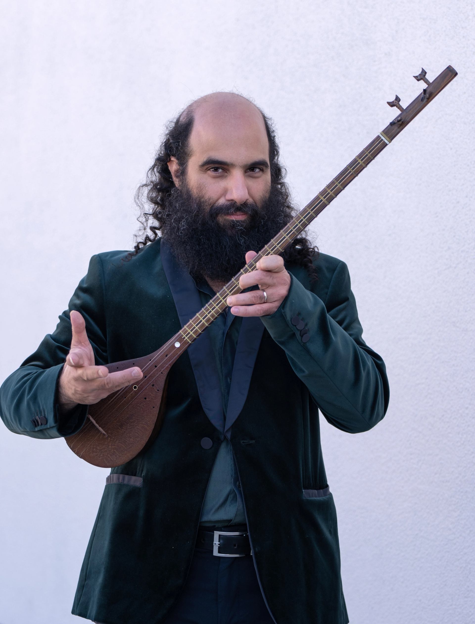 Musician in dark suit holding a long-necked string instrument against a plain wall.
