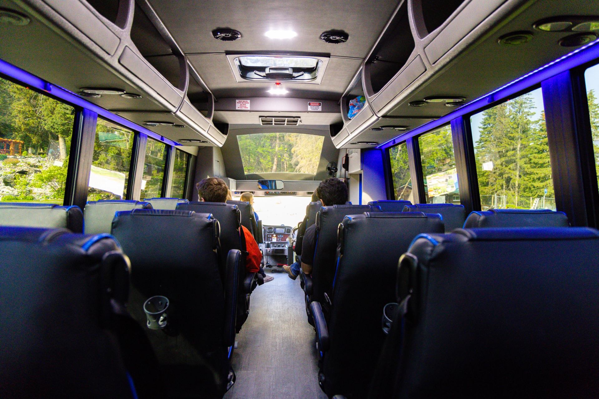 Interior of a coach bus with passengers seated, large windows showing trees outside.