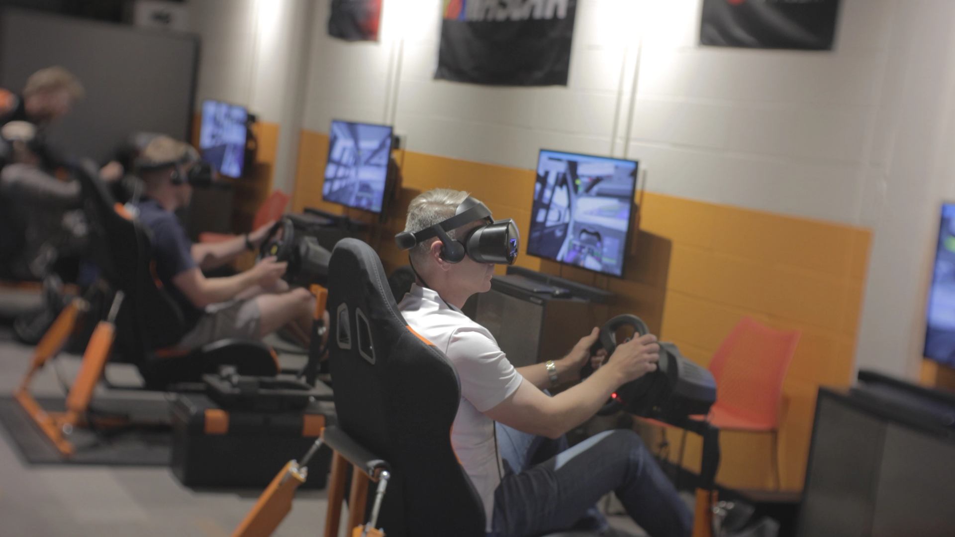 People racing in VR simulators with headsets and steering setups.