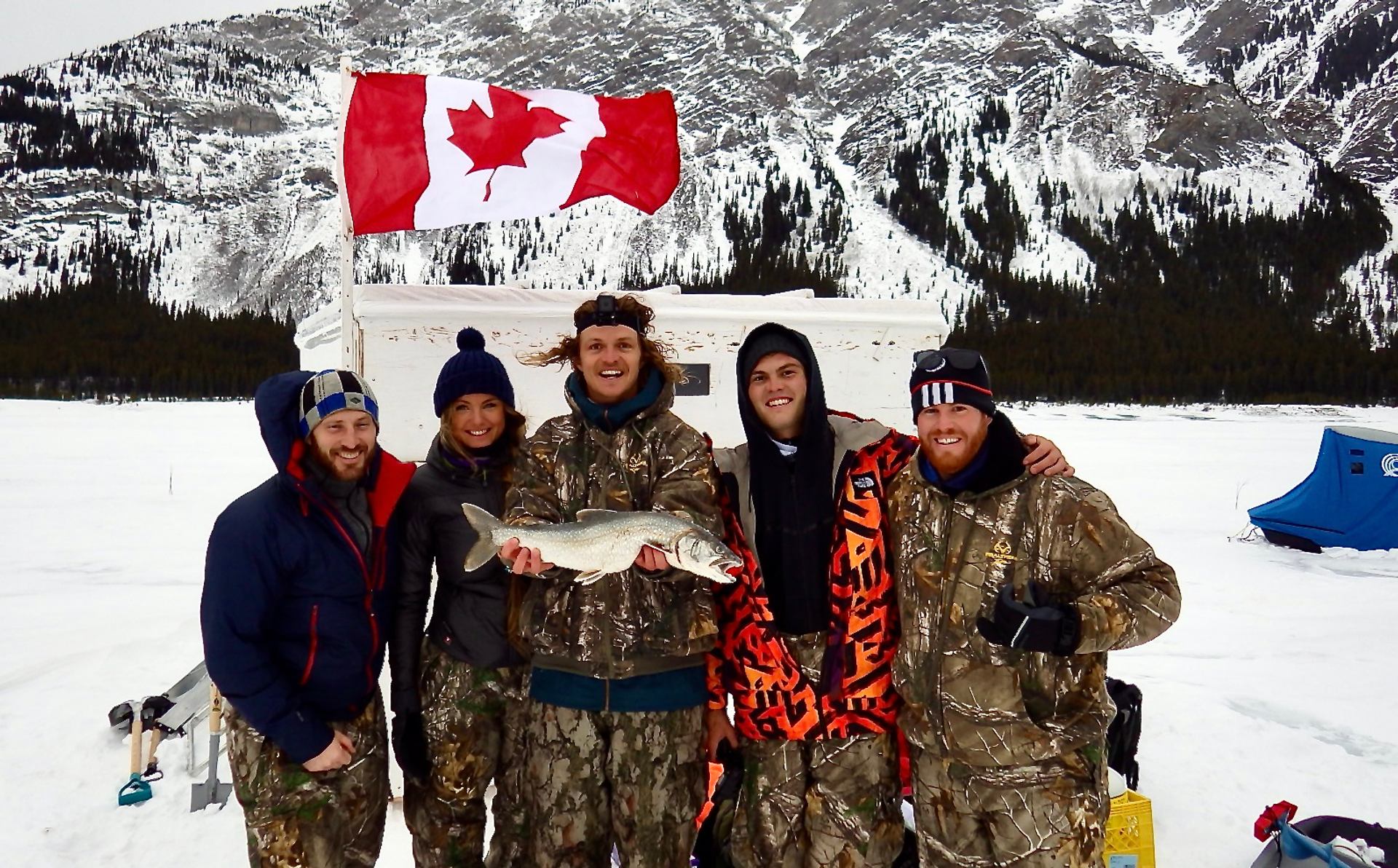 Group of ice fishers holding a fish, Canadian flag and snowy mountains behind.