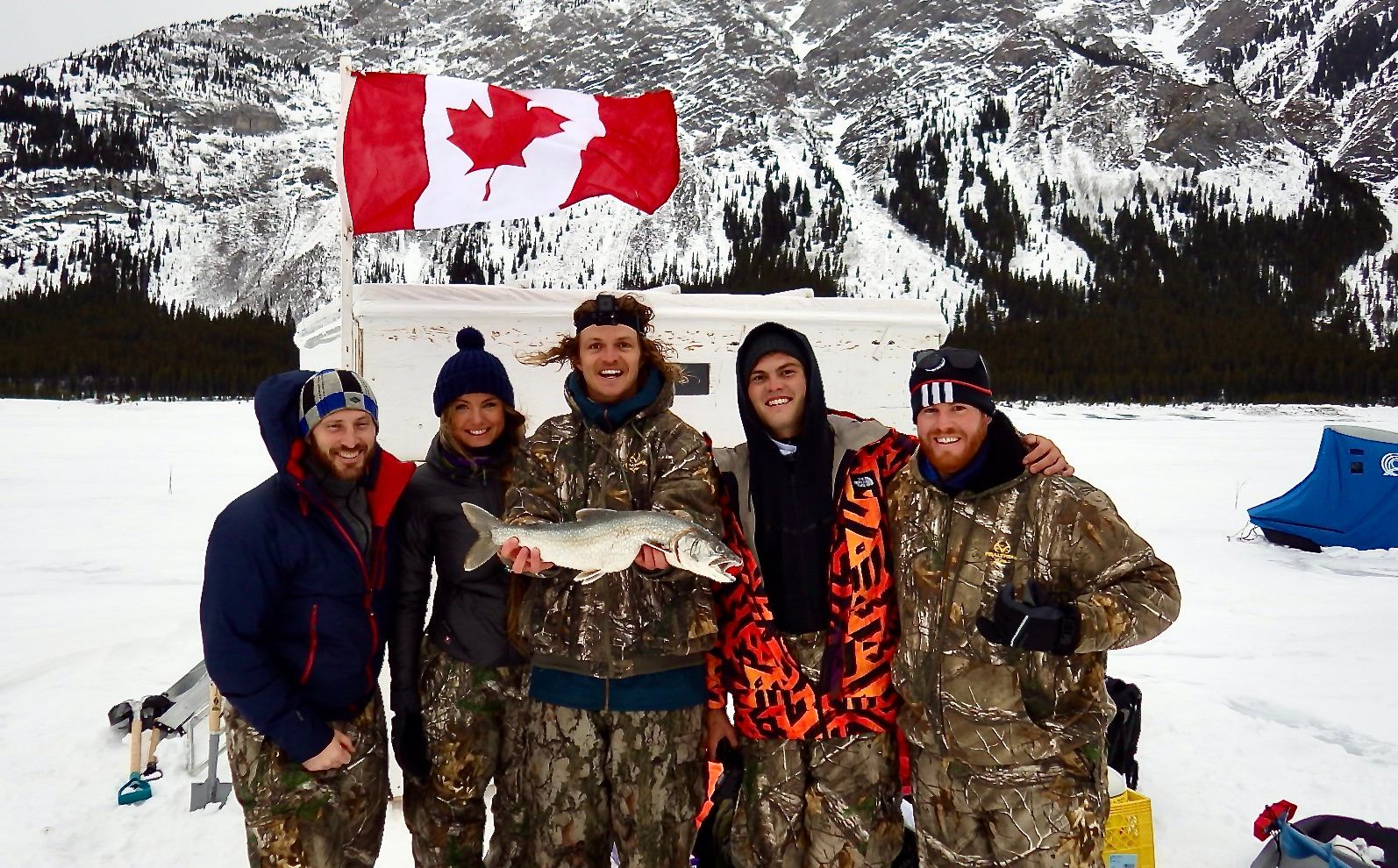 Group of ice fishers holding a fish, Canadian flag and snowy mountains behind.
