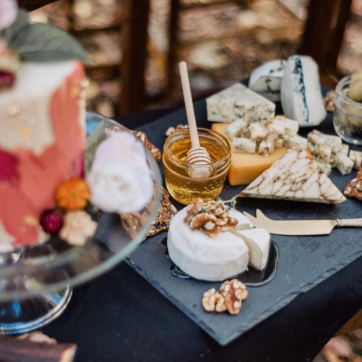 A gourmet cheese platter with various cheeses, walnuts, and a jar of honey with a dipper. A floral-decorated cake sits alongside, creating an elegant setting.