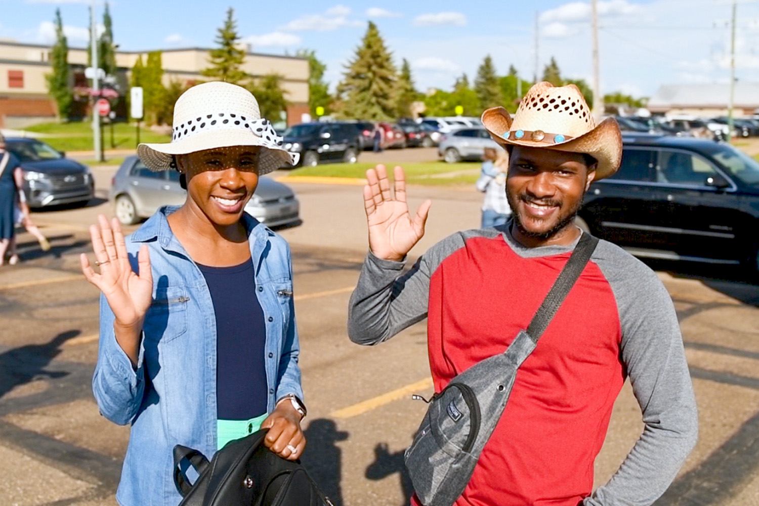 Two people wave in a sunny parking lot with cars, trees, and buildings in the background.
