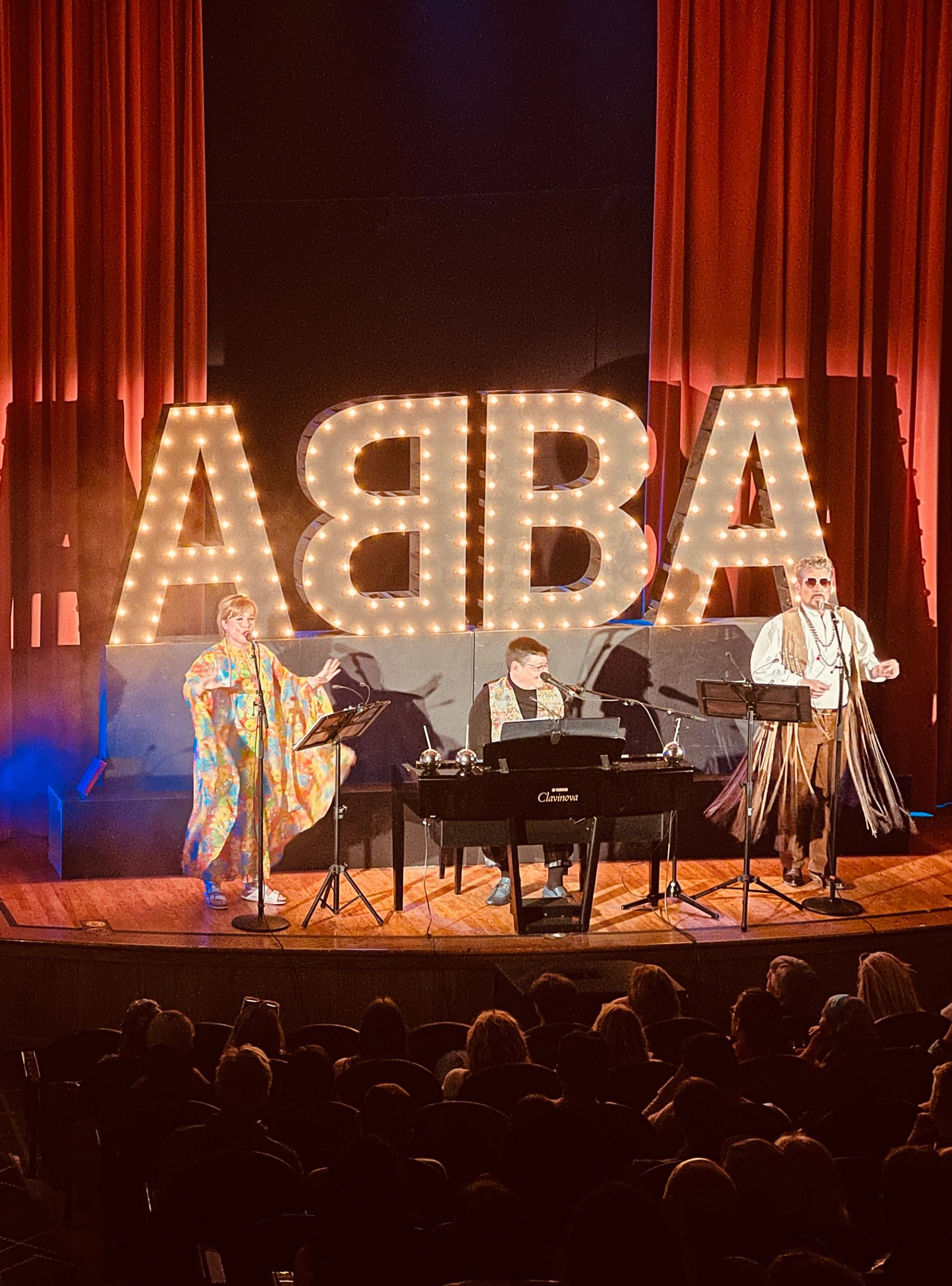 Three performers on stage with piano and glowing “ABBA” letters behind red curtains.