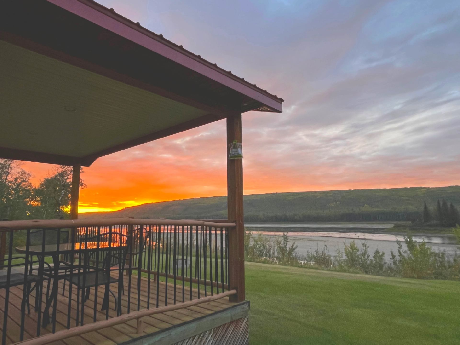 Wooden porch with chairs facing a river and hills under a vibrant orange sunset sky