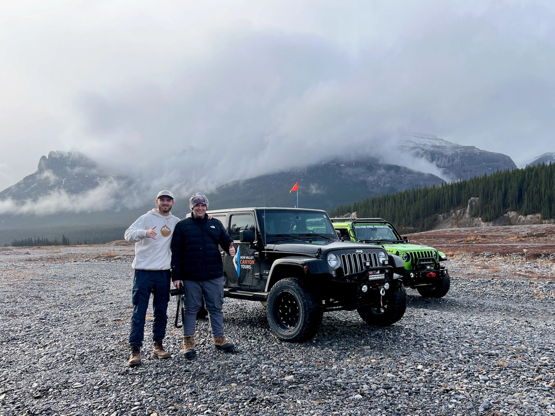 Two people stand beside black and green Jeeps on a rocky trail with misty mountains behind.