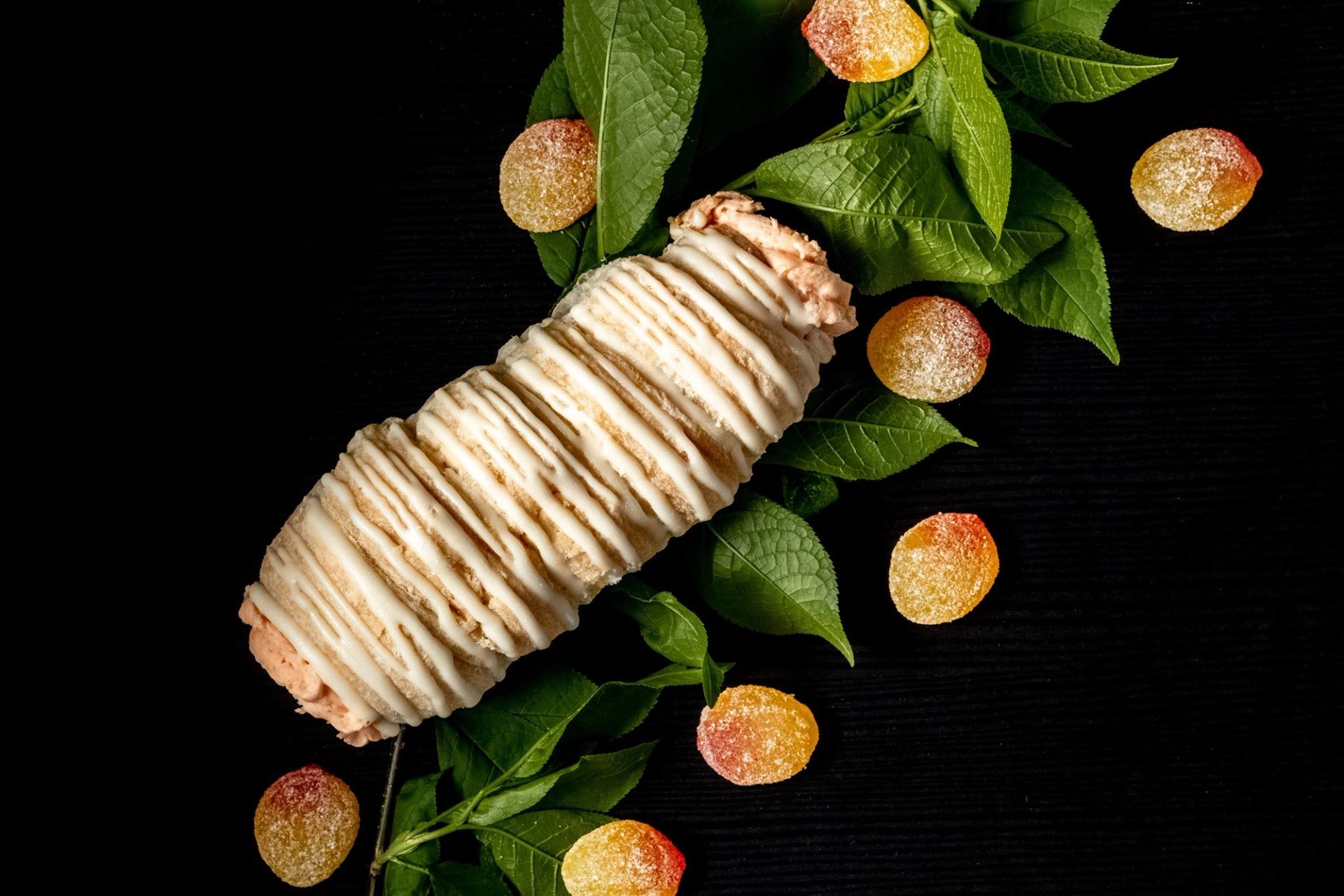 Iced pastry roll on dark surface with green leaves and sugar-coated candies.