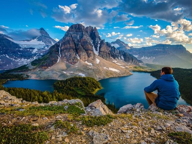 Person sitting on a ridge looking out over blue lakes and dramatic mountain peaks.