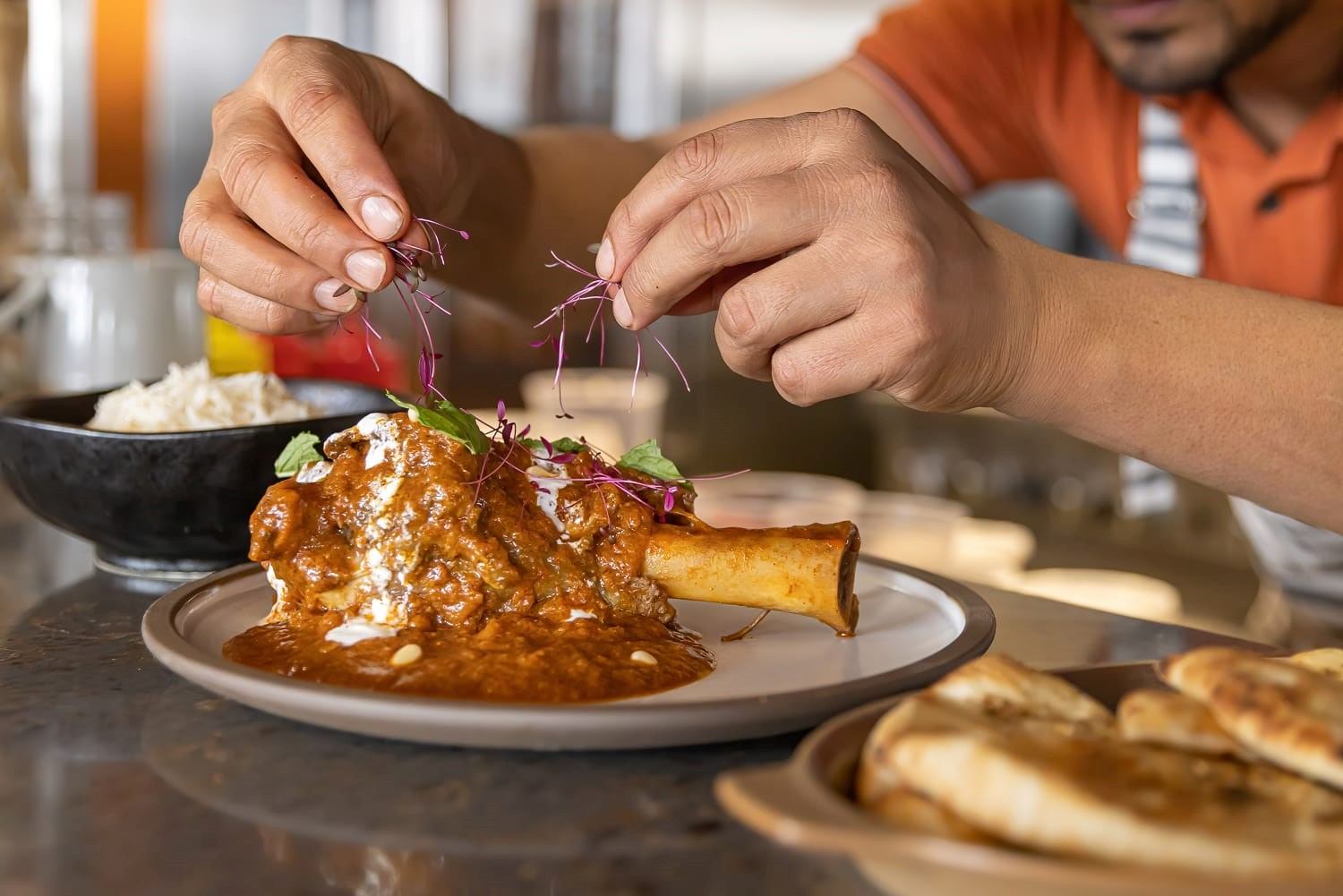 Hands garnish a plated meat dish with microgreens and purple sprouts at a restaurant table.