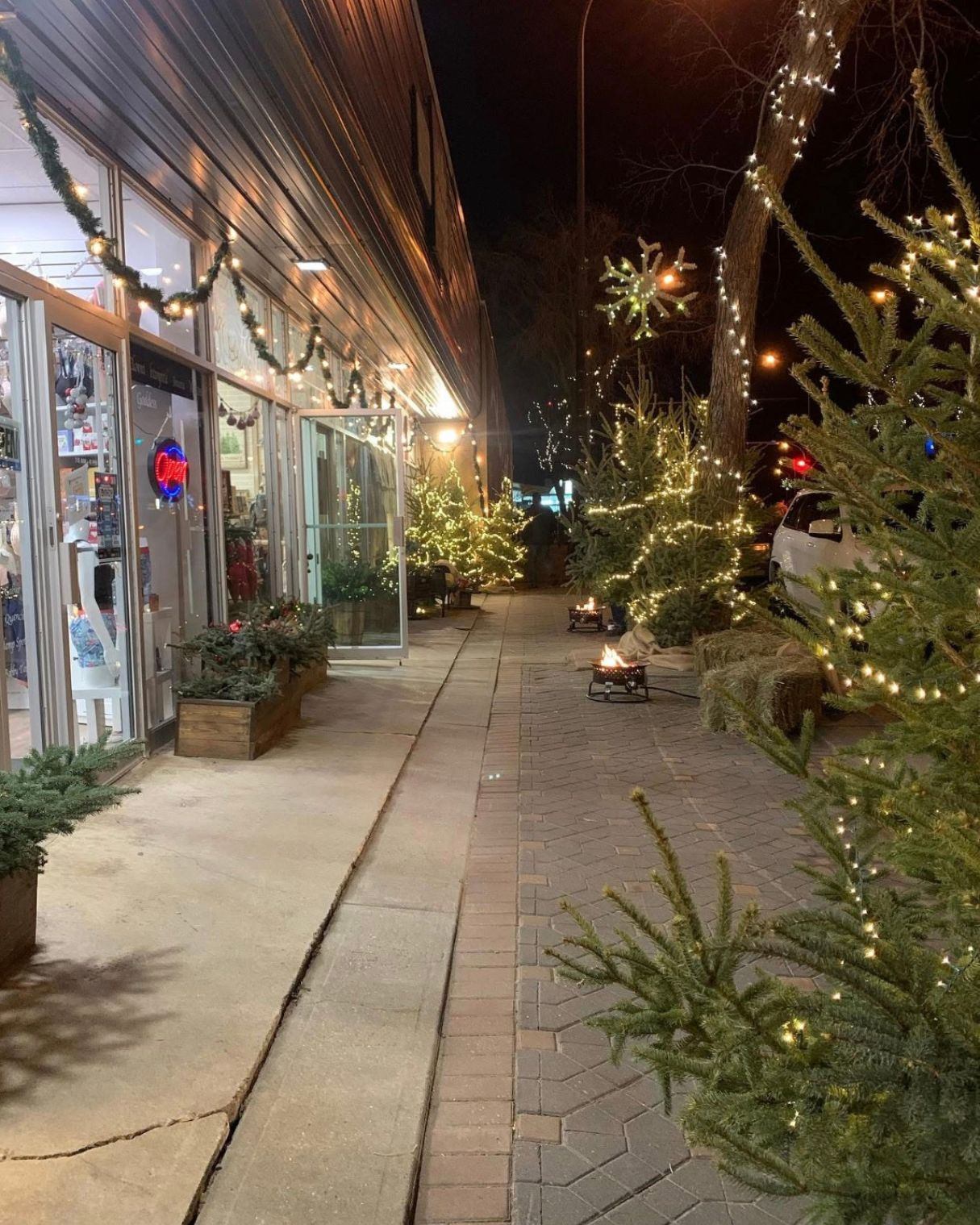 Sidewalk beside shop lined with lit evergreen trees, garlands and small outdoor fire pits.