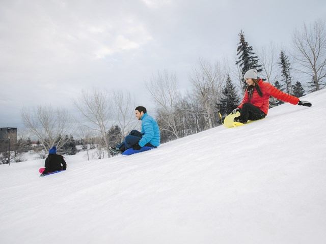 People sitting on a snowy slope with snowboards and skis in a park landscape.