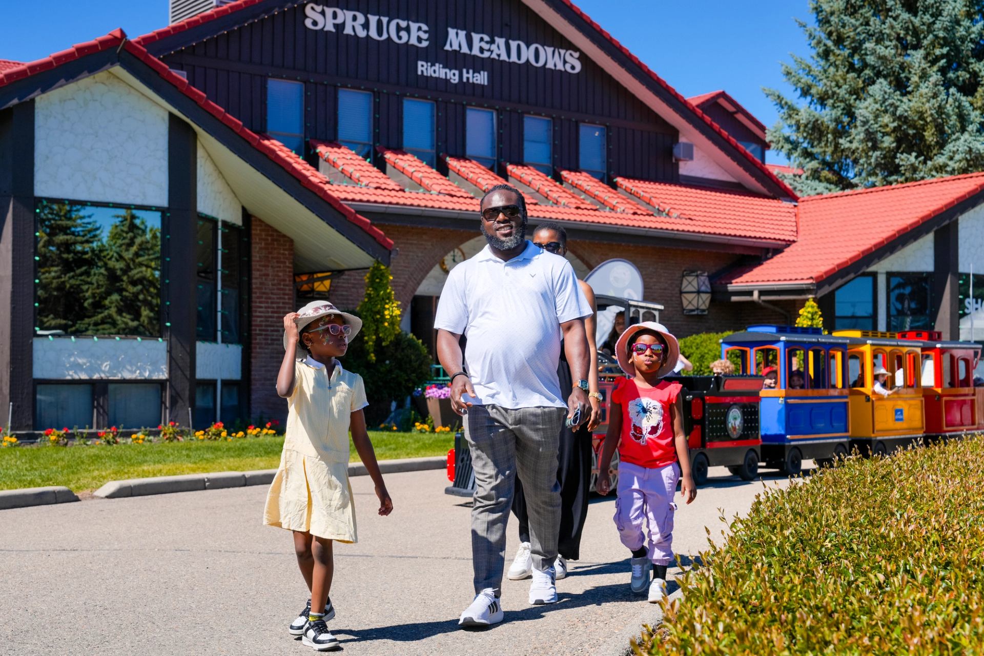 A family walks past the Spruce Meadows Riding Hall with a colorful train behind them.