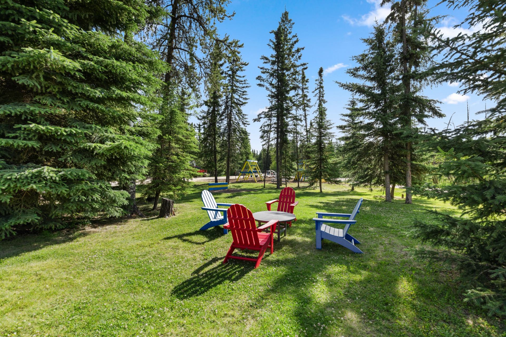 Colorful Adirondack chairs surround a table on a grassy lawn among tall evergreen trees.