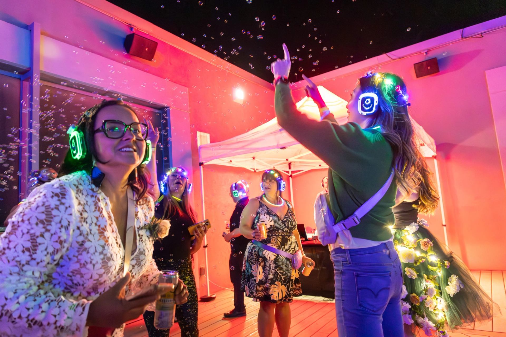 People wearing glowing headphones dance under bubbles and colourful lights near a white canopy.