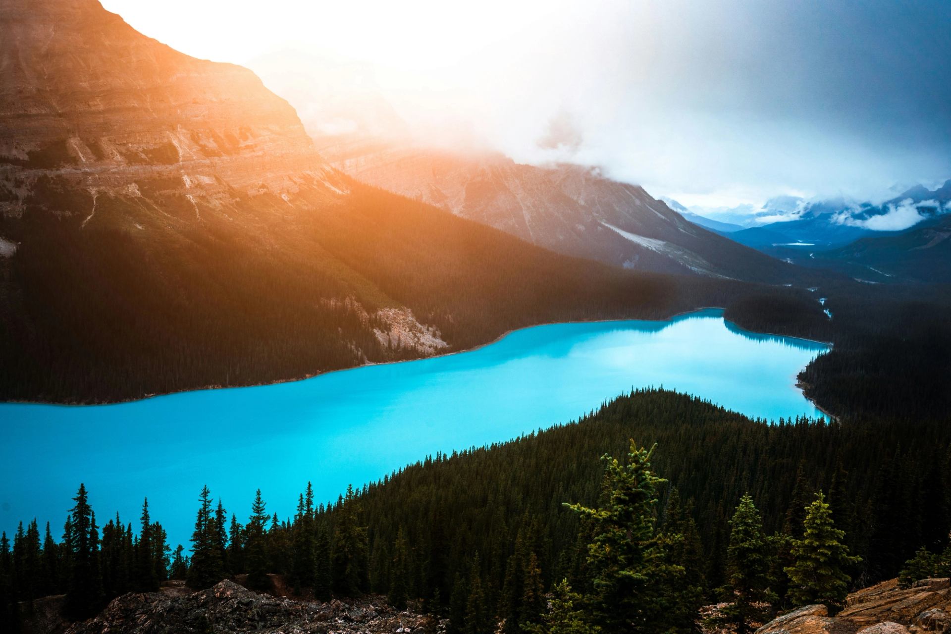 Turquoise mountain lake curves through dense evergreen forest under hazy sunlit peaks.