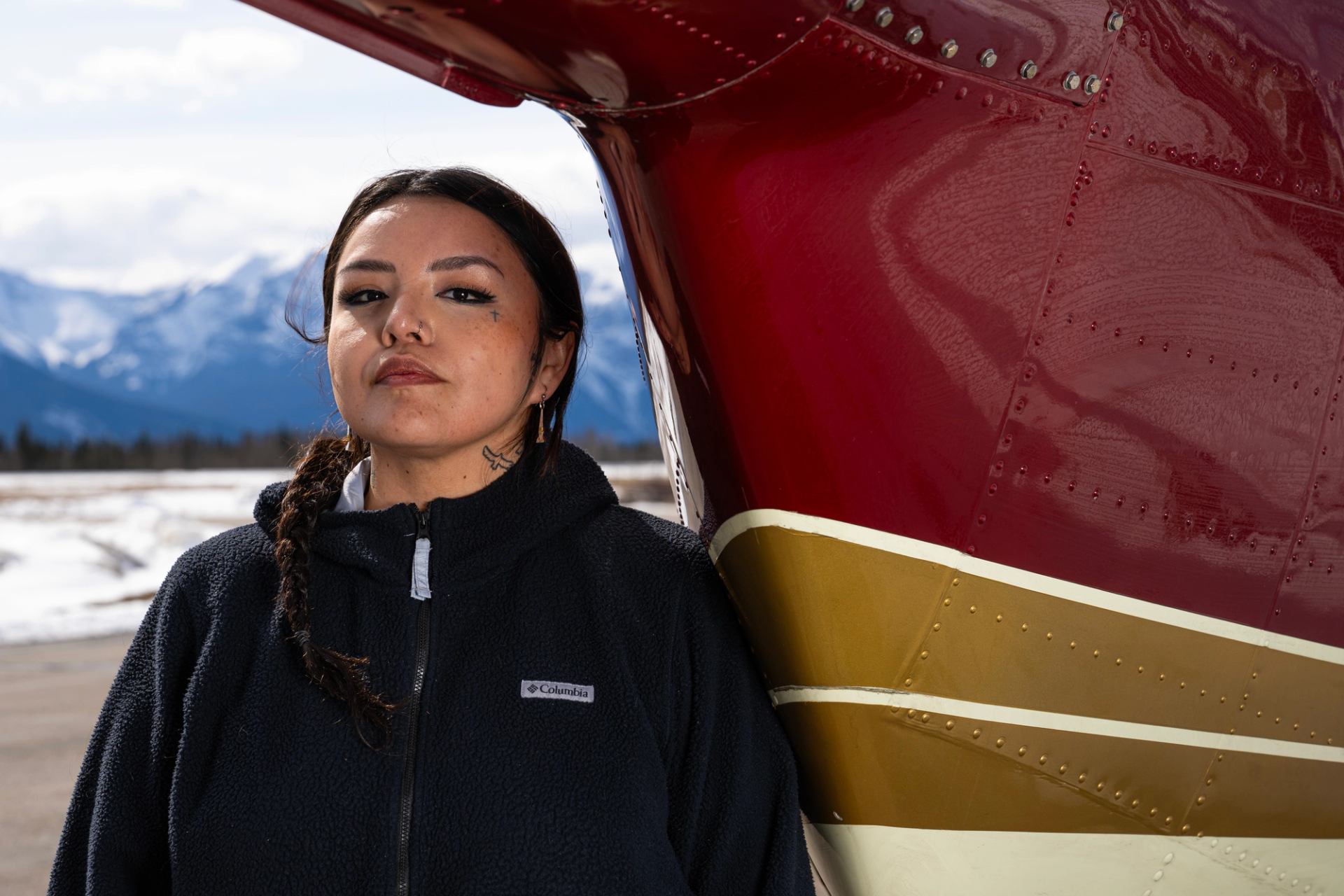 Person stands beside red and yellow aircraft with snowy mountains in the background.