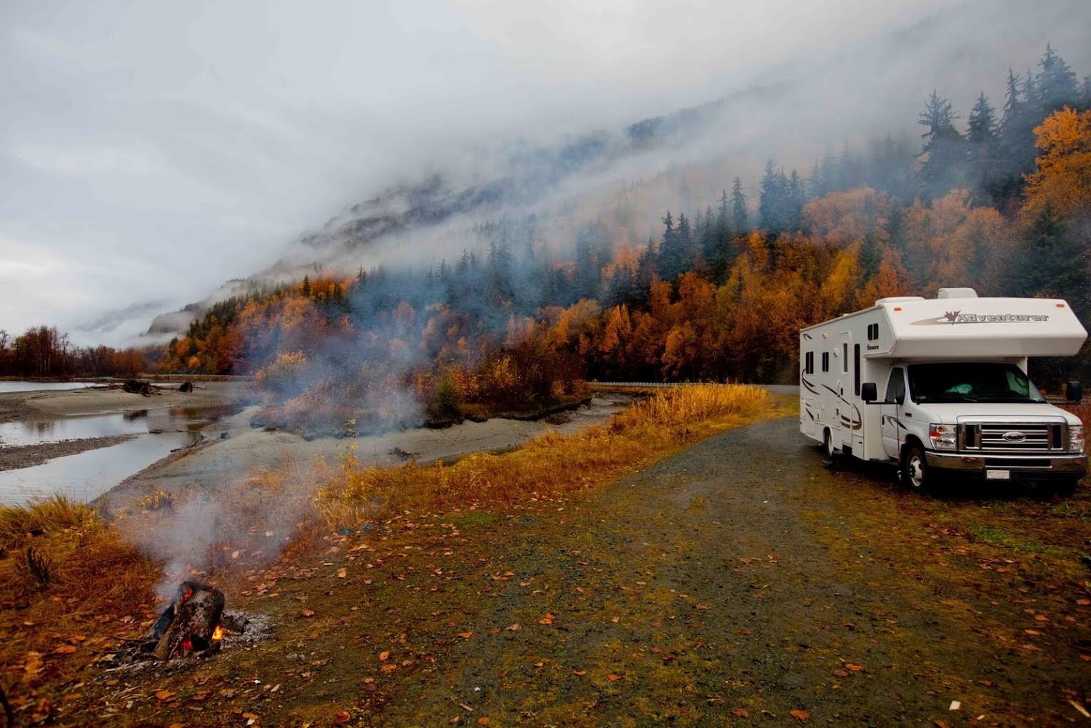 Fraserway RV parked near campfire in misty autumn forest with mountain backdrop.