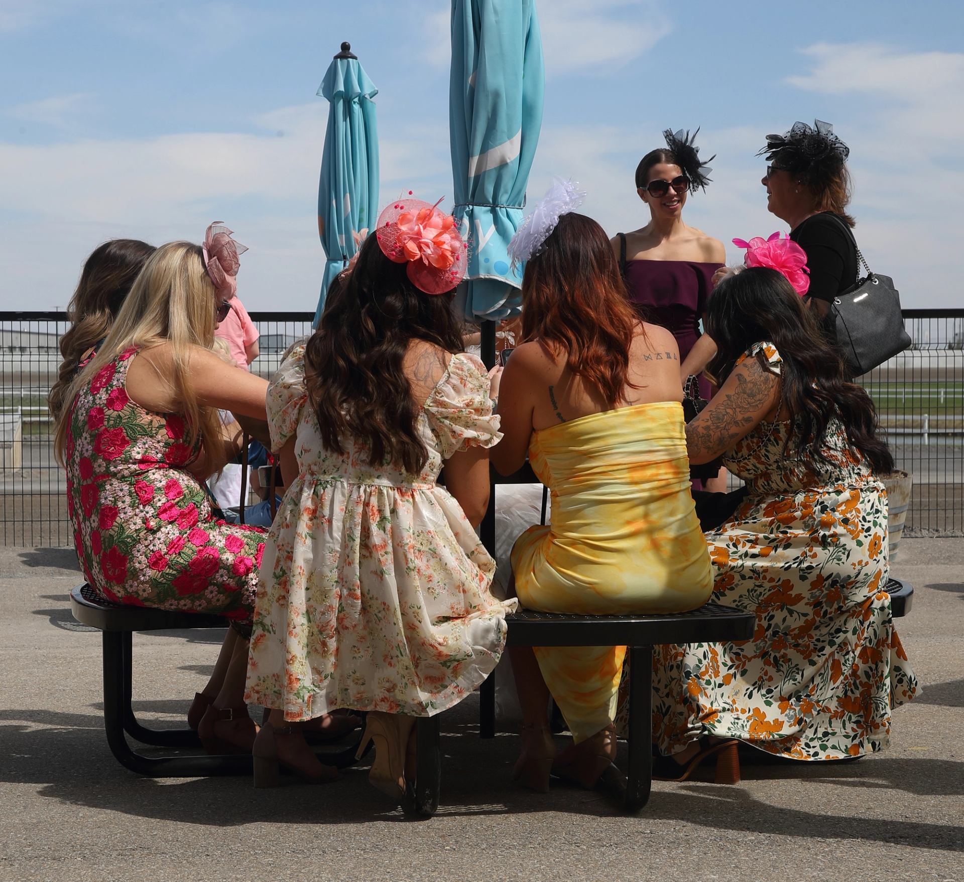 Group of people in colorful dresses and hats seated at an outdoor table near a racetrack.