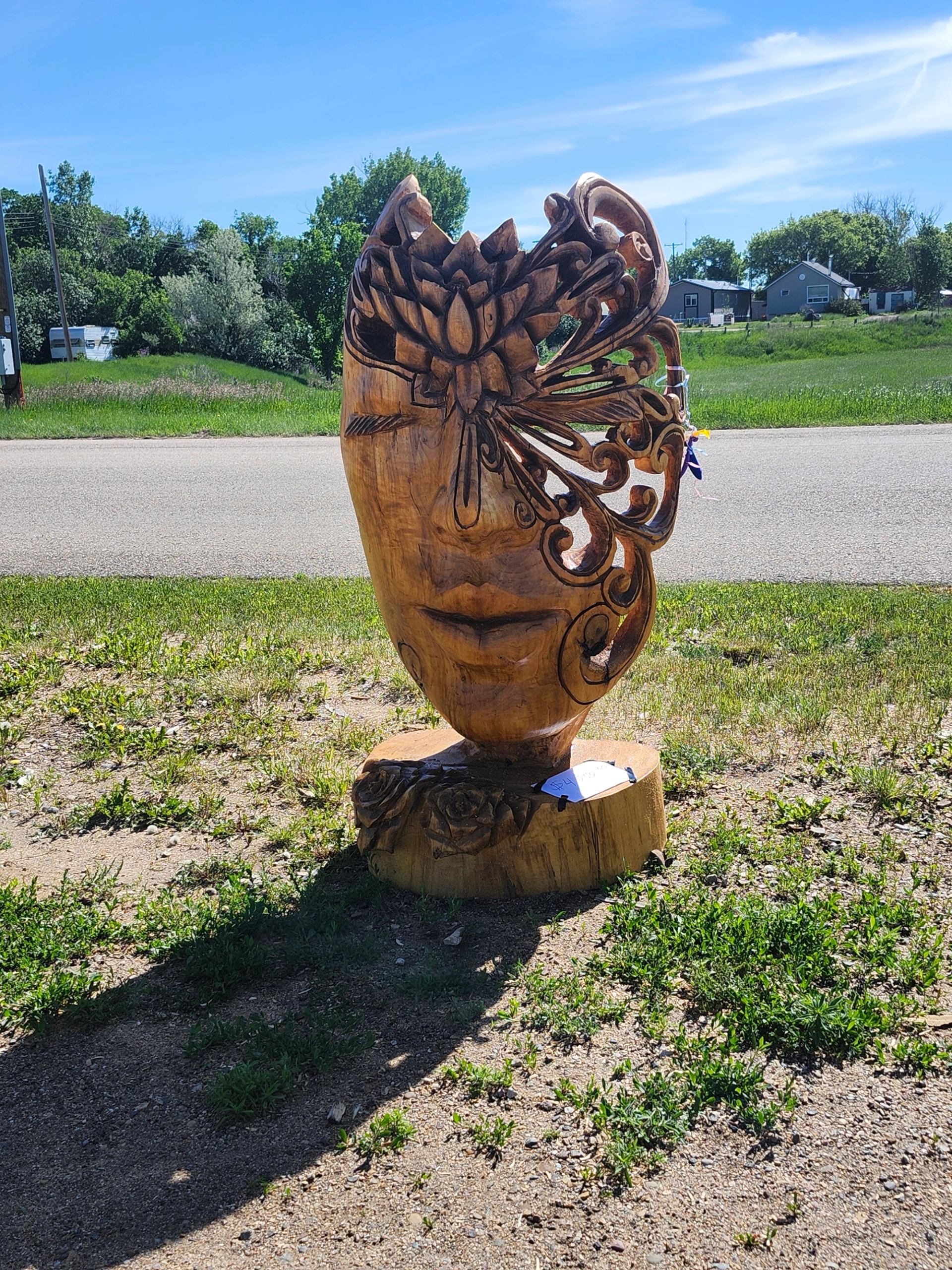 Wooden face sculpture with floral carvings stands beside rural road and grassy field.