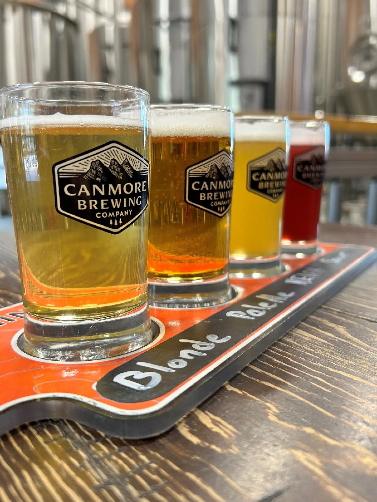 Flight of Canmore Brewing beers on wooden table with brewery tanks in background.