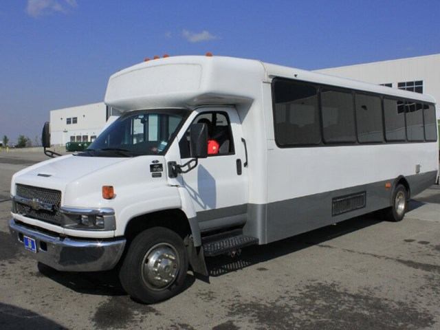 White CST Transportations bus with large windows parked in front of building.