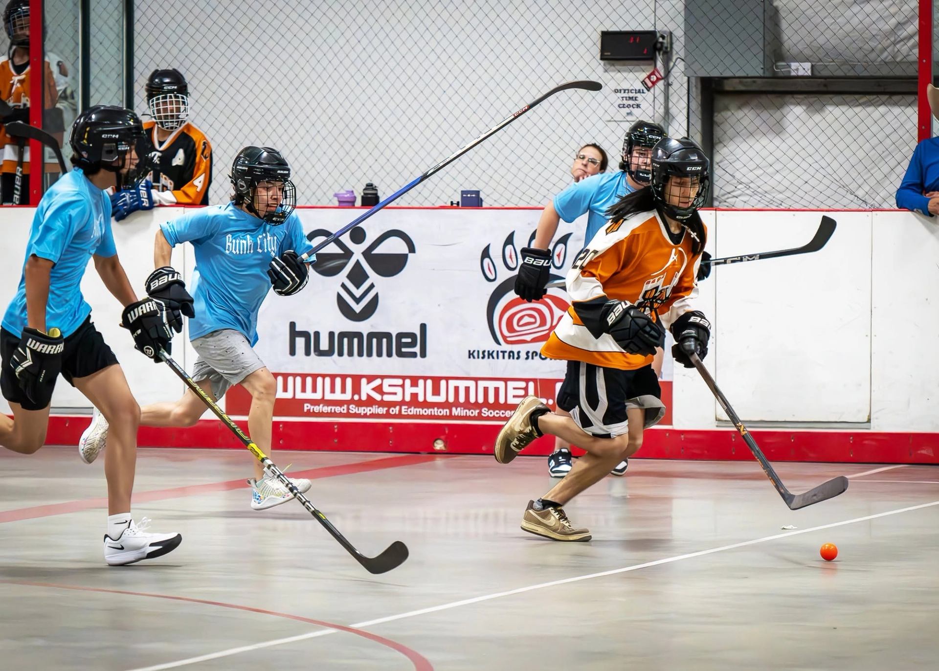 Ball hockey players in blue and orange jerseys chase an orange ball indoors.