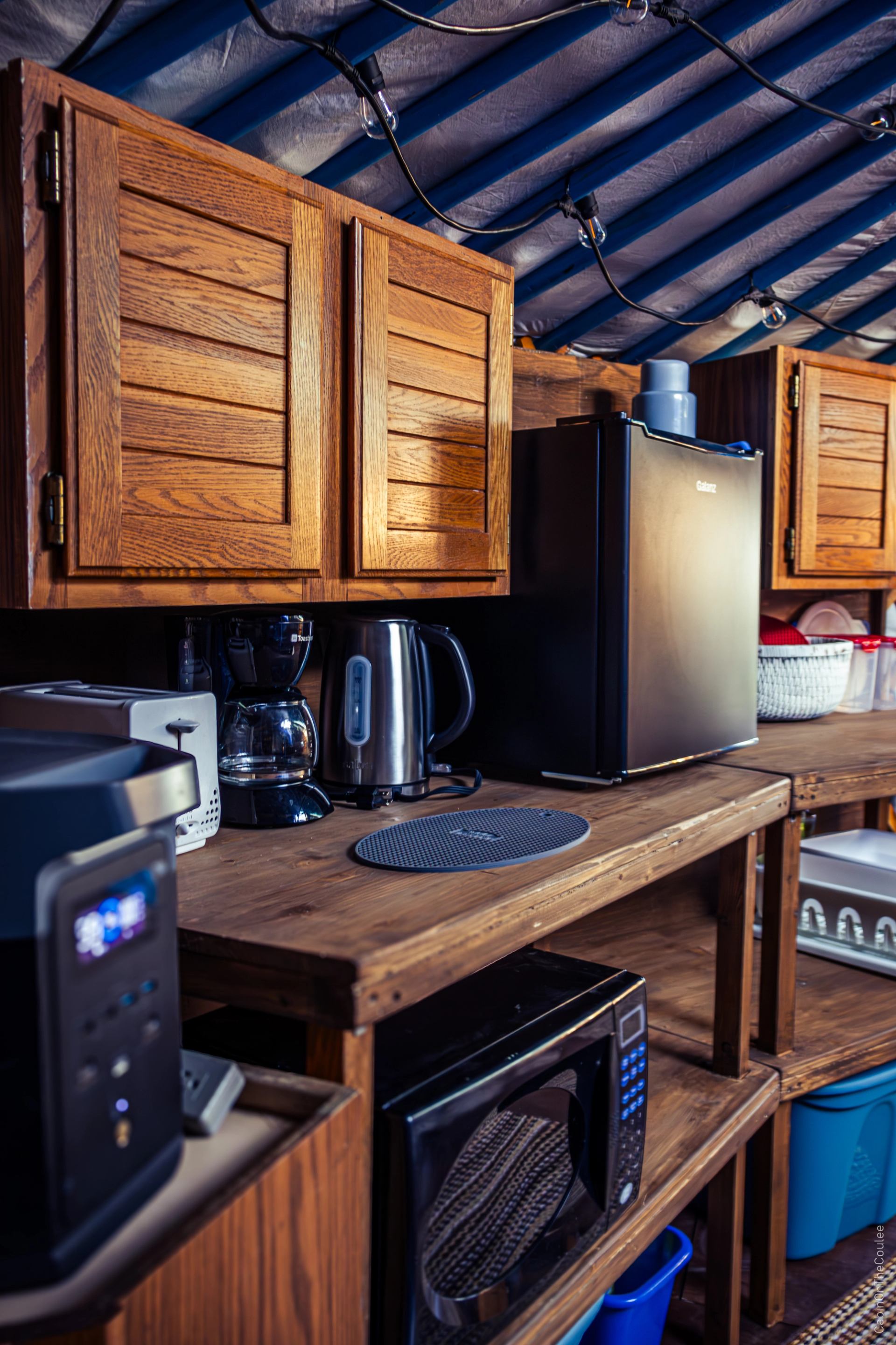 Wooden kitchenette with fridge, microwave, kettle, coffee maker, and cabinets.