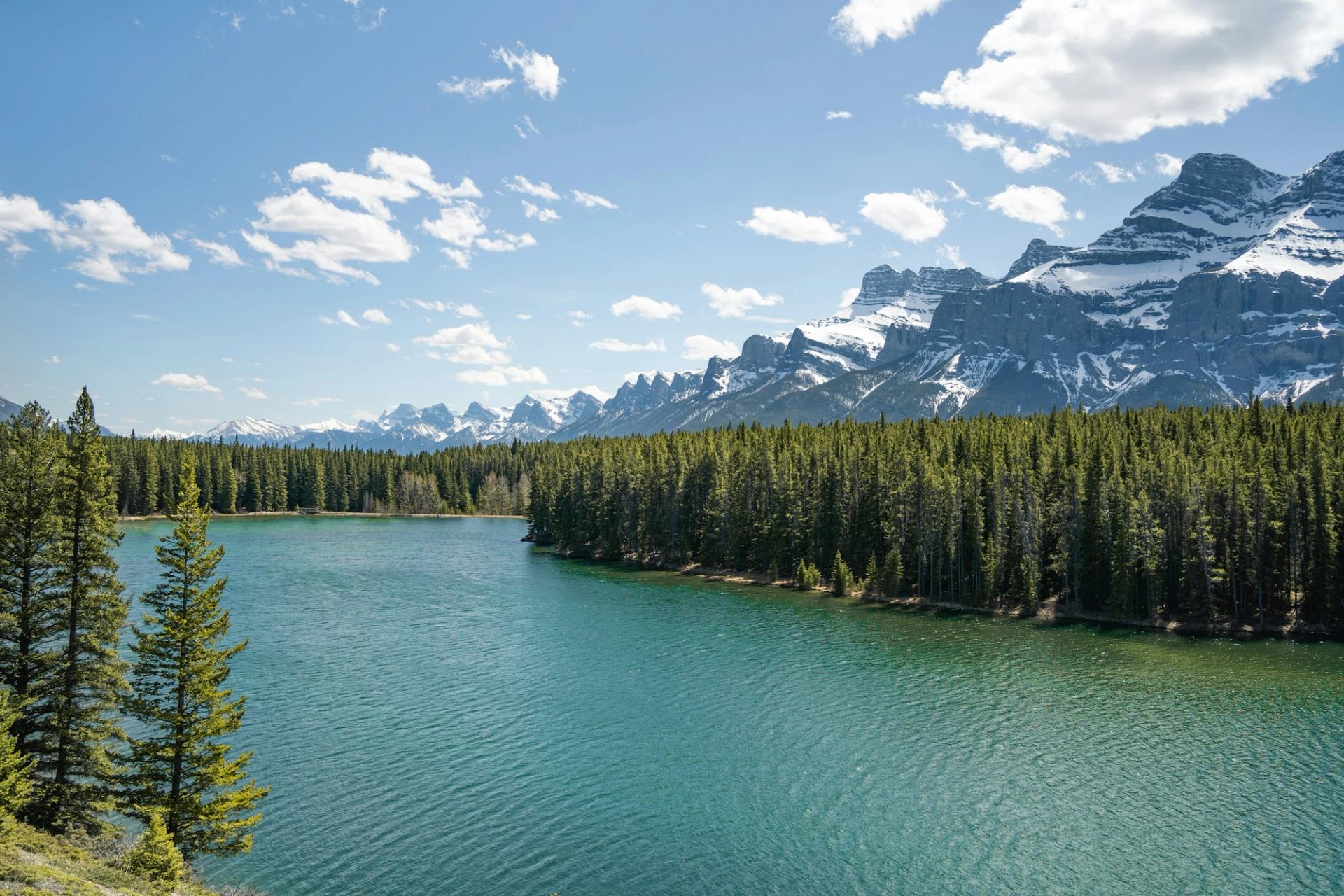 Clear turquoise lake surrounded by dense pine forest and rugged snow-capped peaks.