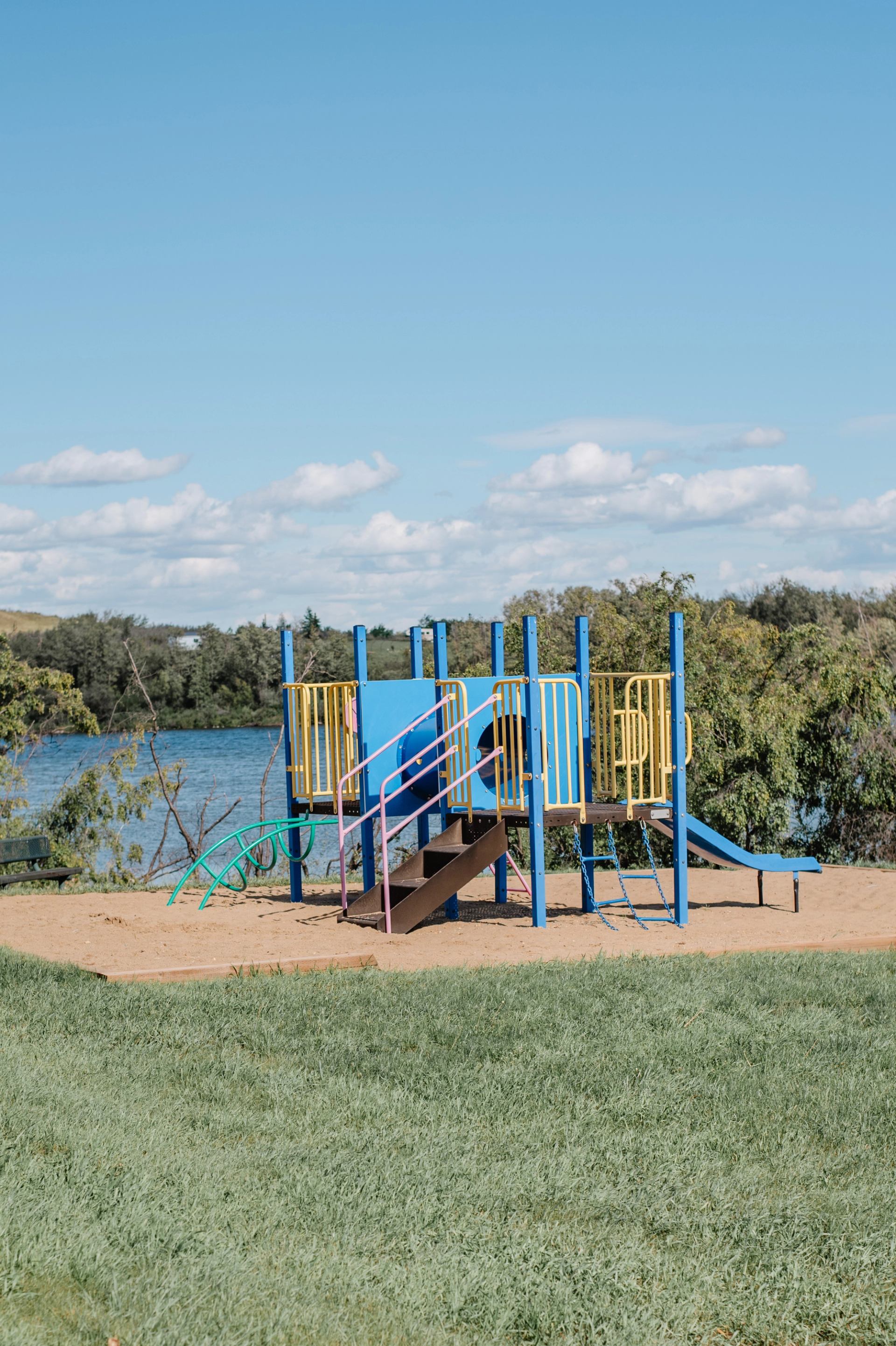 Playground structure with slide and stairs on sand beside a lake and trees.