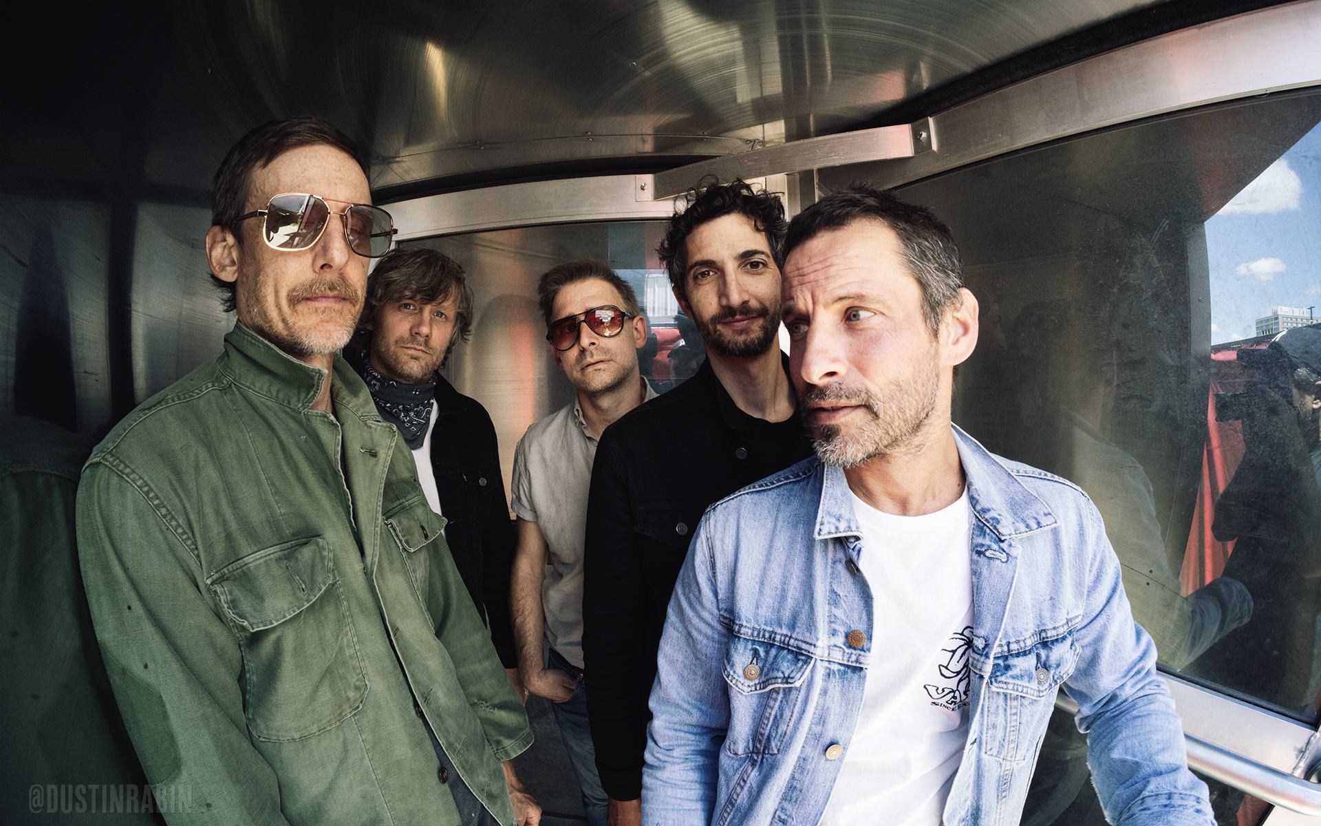 Five men in casual attire and sunglasses pose together in a sleek, metallic interior with a large window.