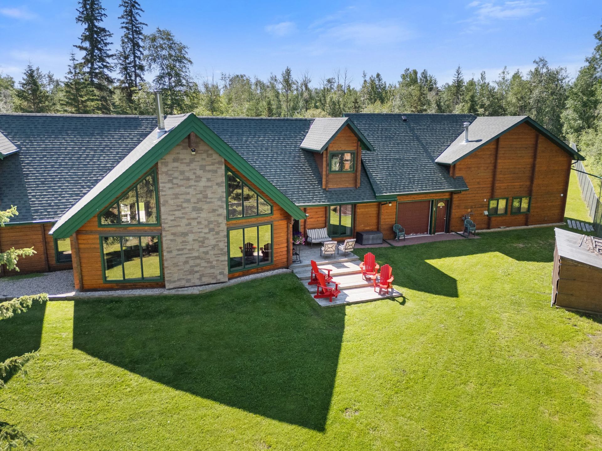 Large wood-sided house with big windows, patio seating, and lawn surrounded by trees.