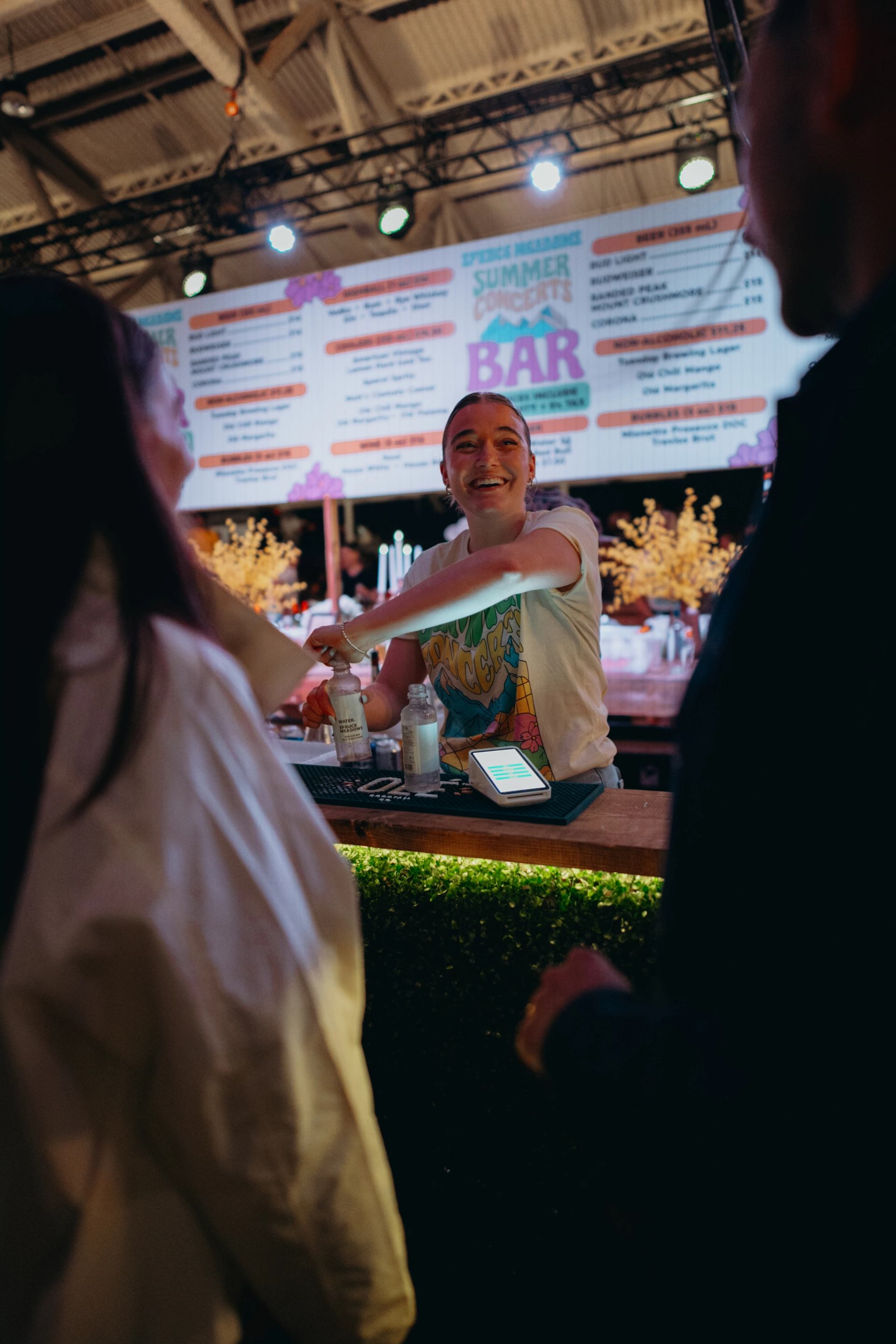A smiling bartender serves drinks to customers at an outdoor bar with a menu overhead.
