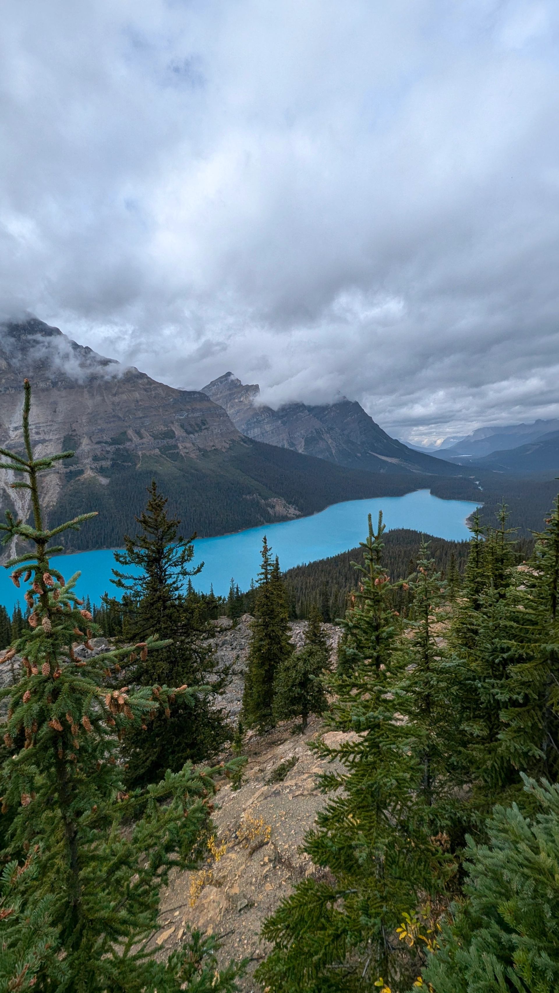 Turquoise lake surrounded by pine trees and rugged mountains under cloudy skies.