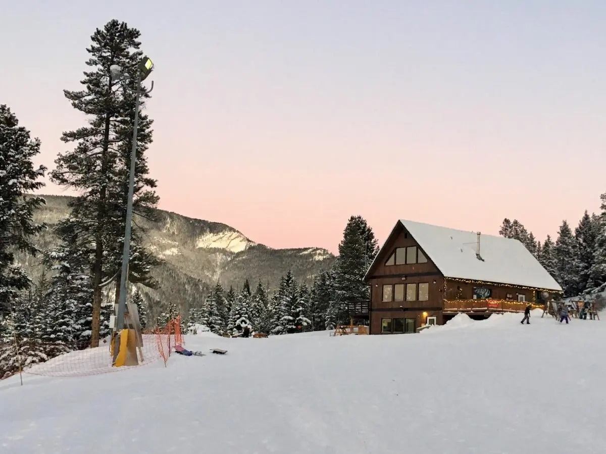 Snow-covered lodge at Pass Powderkeg with mountains and trees at sunset.
