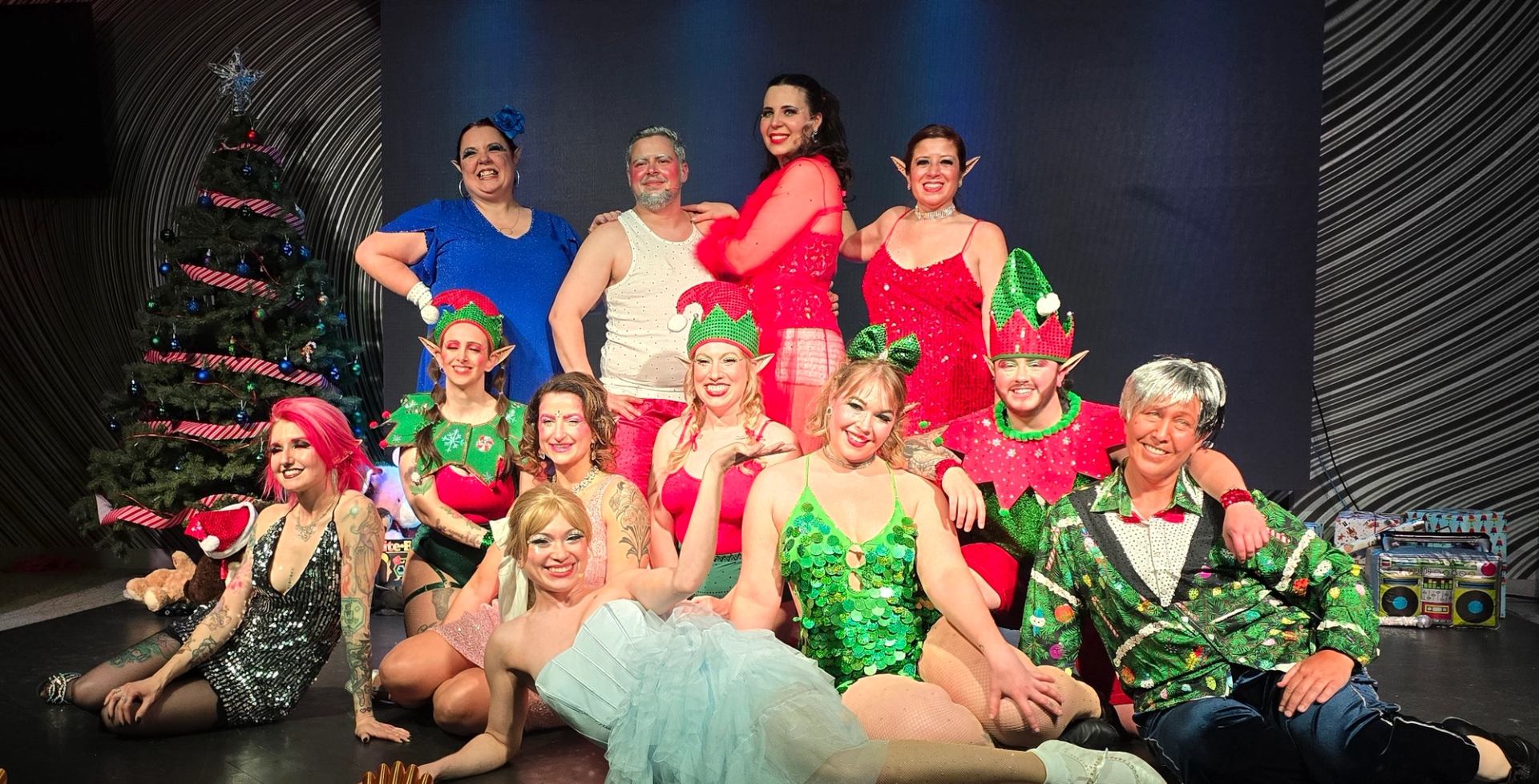 Group of performers in festive costumes posing onstage beside decorated Christmas tree.