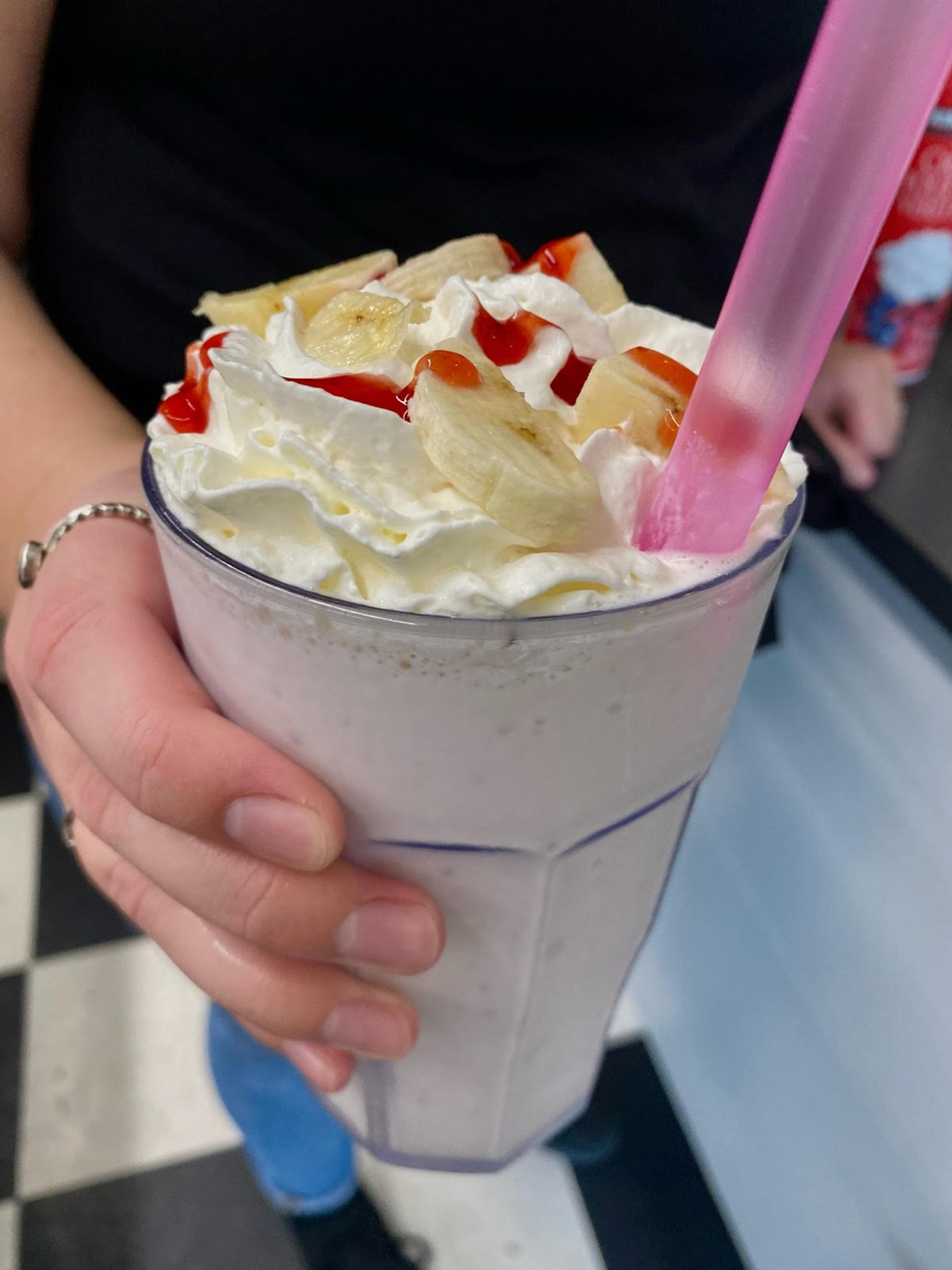 Hand holding tall milkshake with whipped cream, banana slices, red syrup and pink straw.