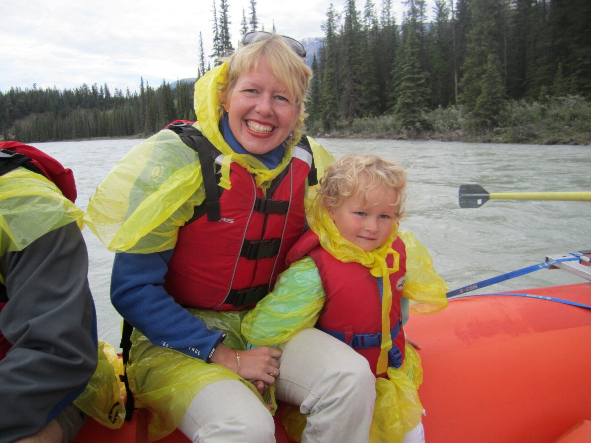 An adult and child smile in rain ponchos while seated in a raft on the river.