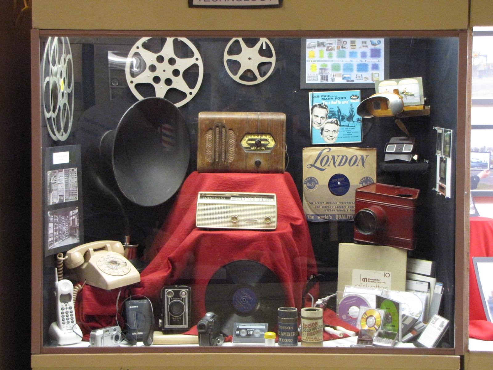 Museum display of vintage radios, records, telephones, and film reels arranged in a glass case.