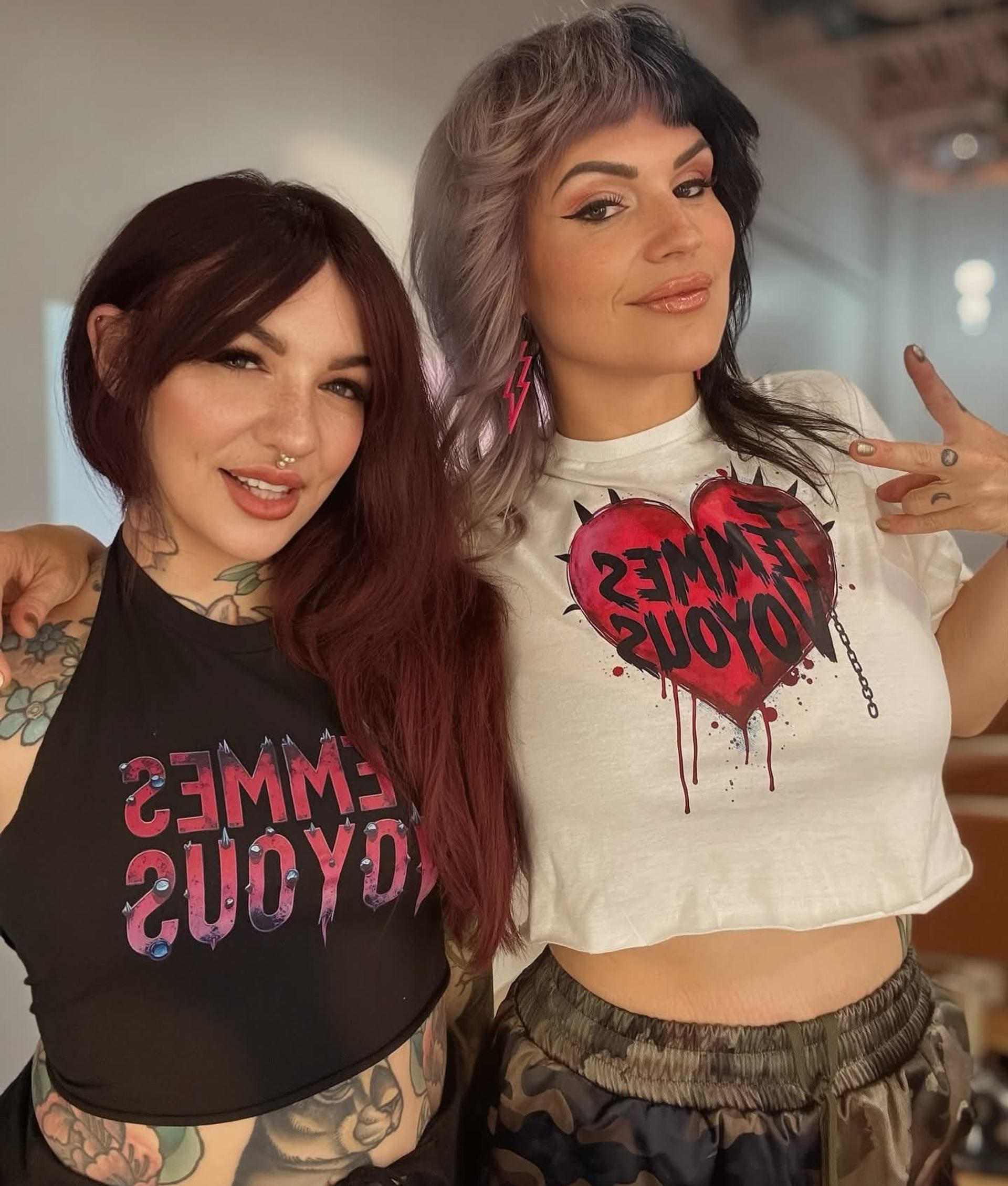 Two tattooed people pose together in graphic tops, one making a hand gesture.