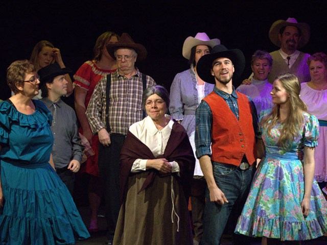 Stage cast in cowboy hats performing in costumes under purple and blue lights.
