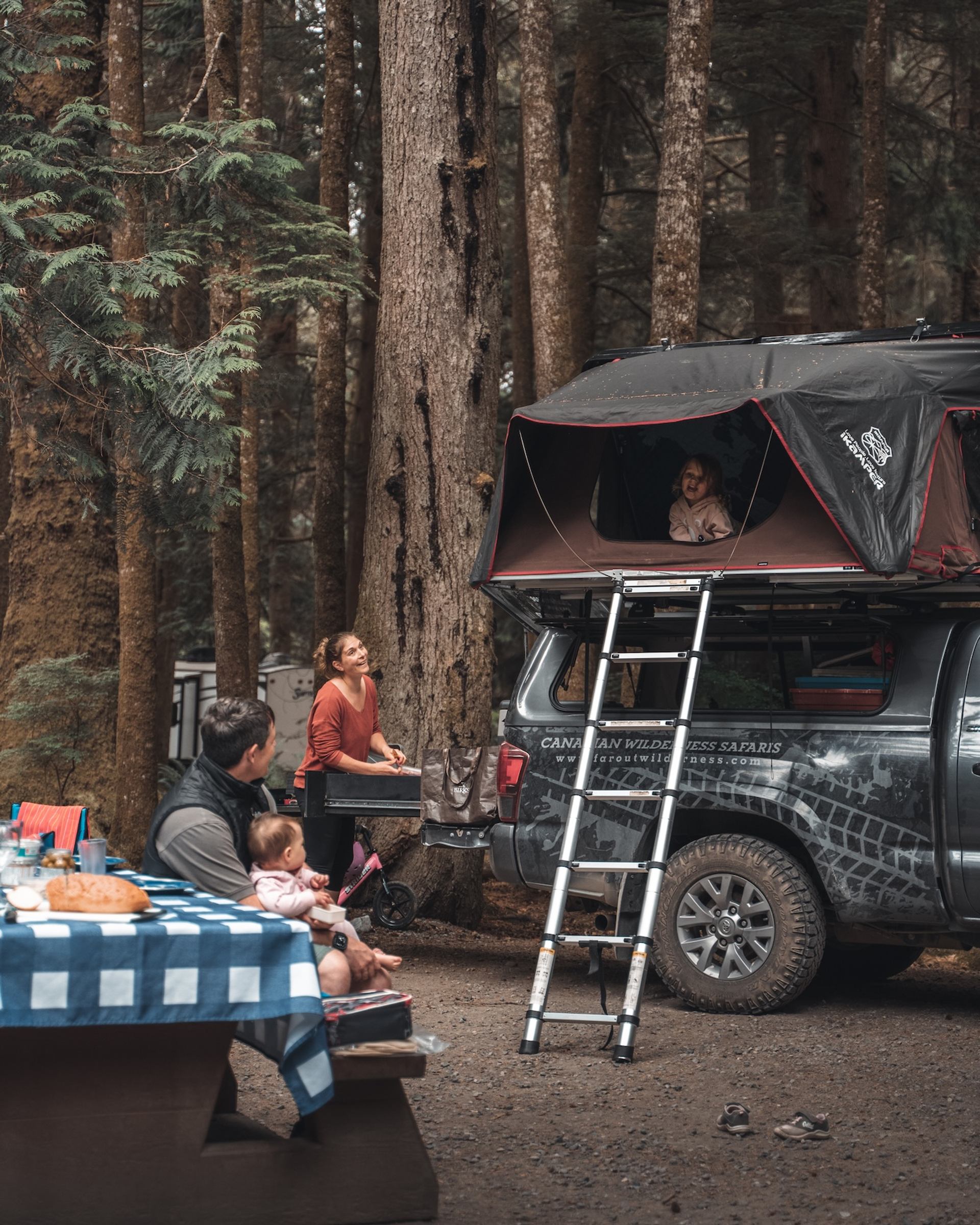 Truck with rooftop tent and family camping in a forest surrounded by tall trees.